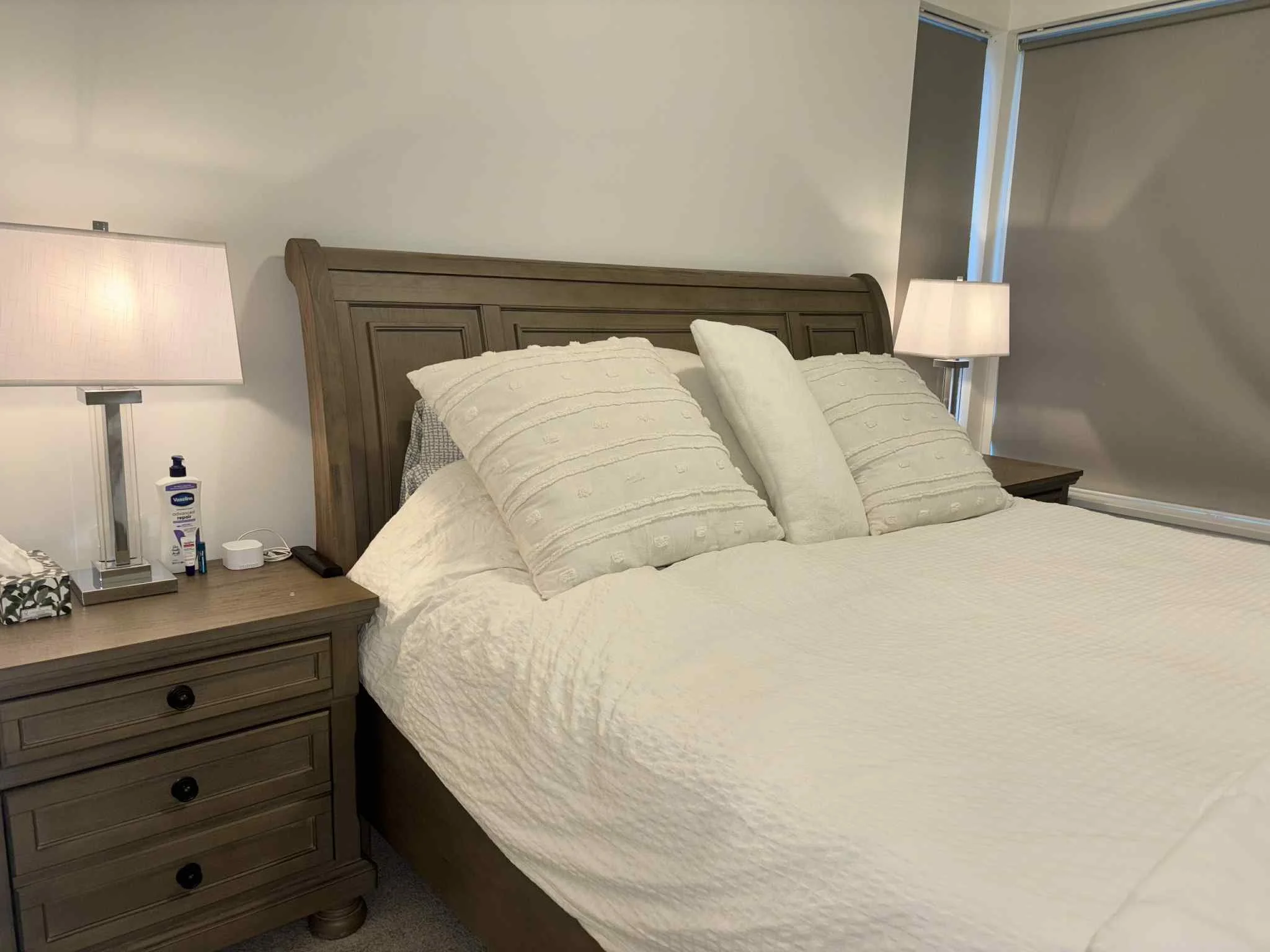 A neatly made bed with a wooden headboard, white quilt, three decorative pillows, two bedside tables with lamps, lotion, and other small items in a bedroom.