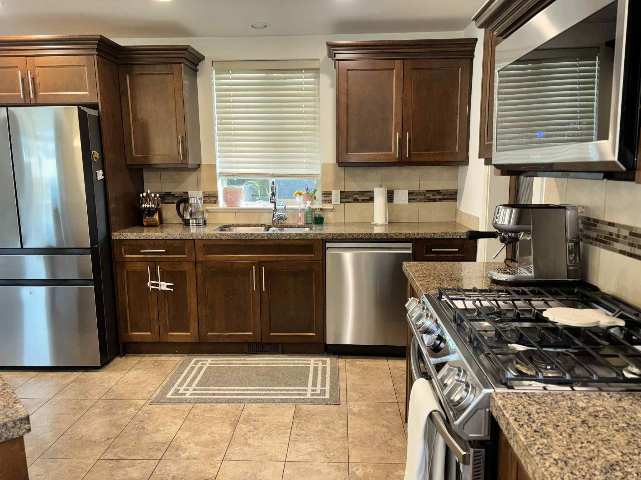 Kitchen with wooden cabinets, stainless steel refrigerator, microwave, oven with stove, granite countertops, tile flooring, window with blinds, coffee maker, sink, paper towels, and various kitchen items.