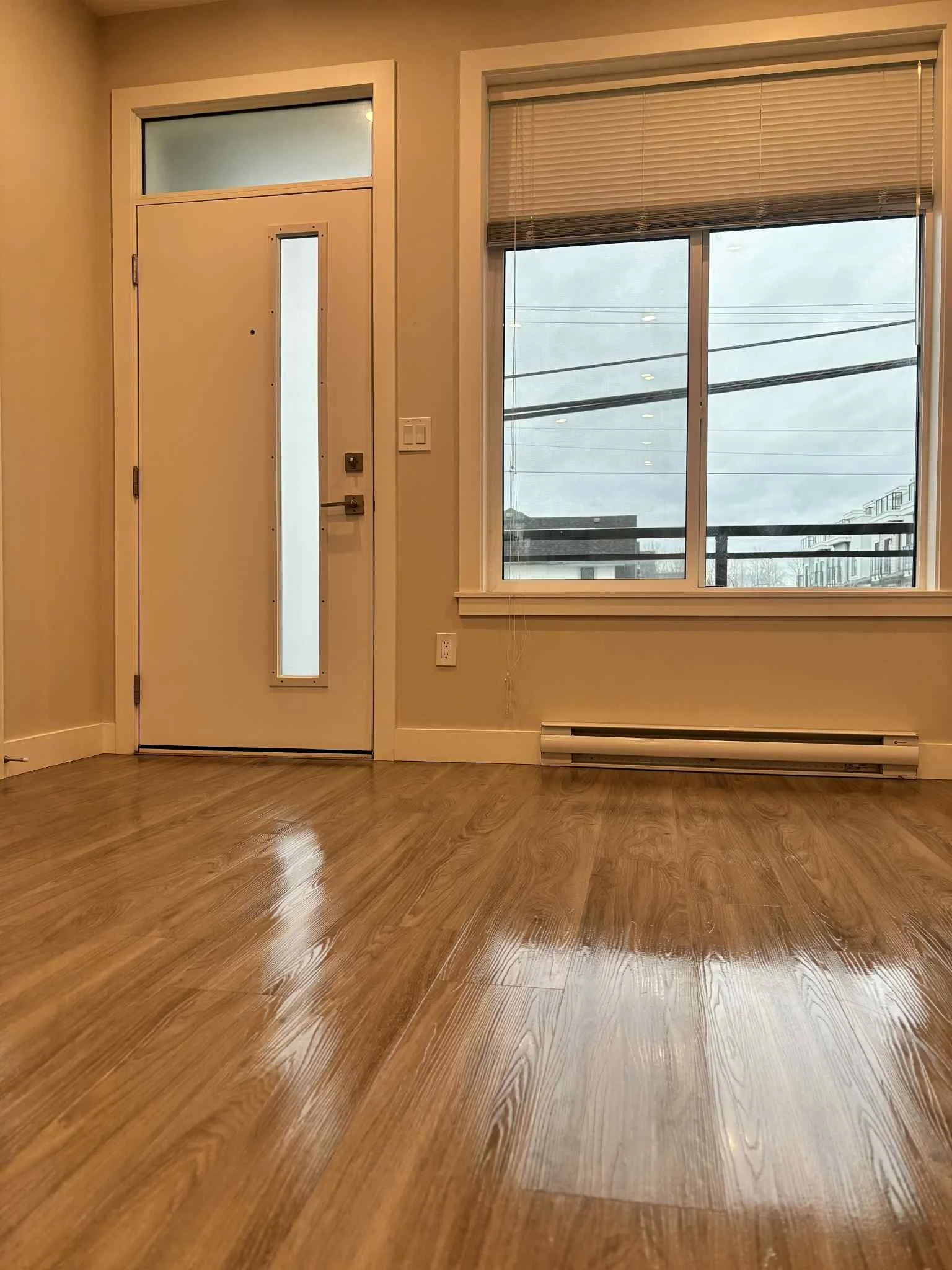 Empty room with wooden flooring, a window with blinds, and a door with a narrow window, some light switches, and a heater along the wall.
