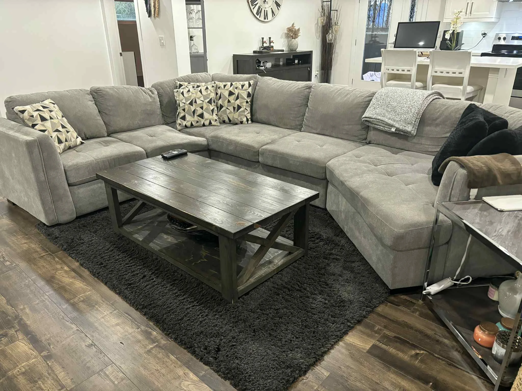 A large gray sectional sofa with patterned and solid black cushions, a gray blanket, and a brown throw blanket, positioned on a black rug in a living room with wood flooring. A wooden coffee table with a dark finish is in front of the sofa, and a rem
