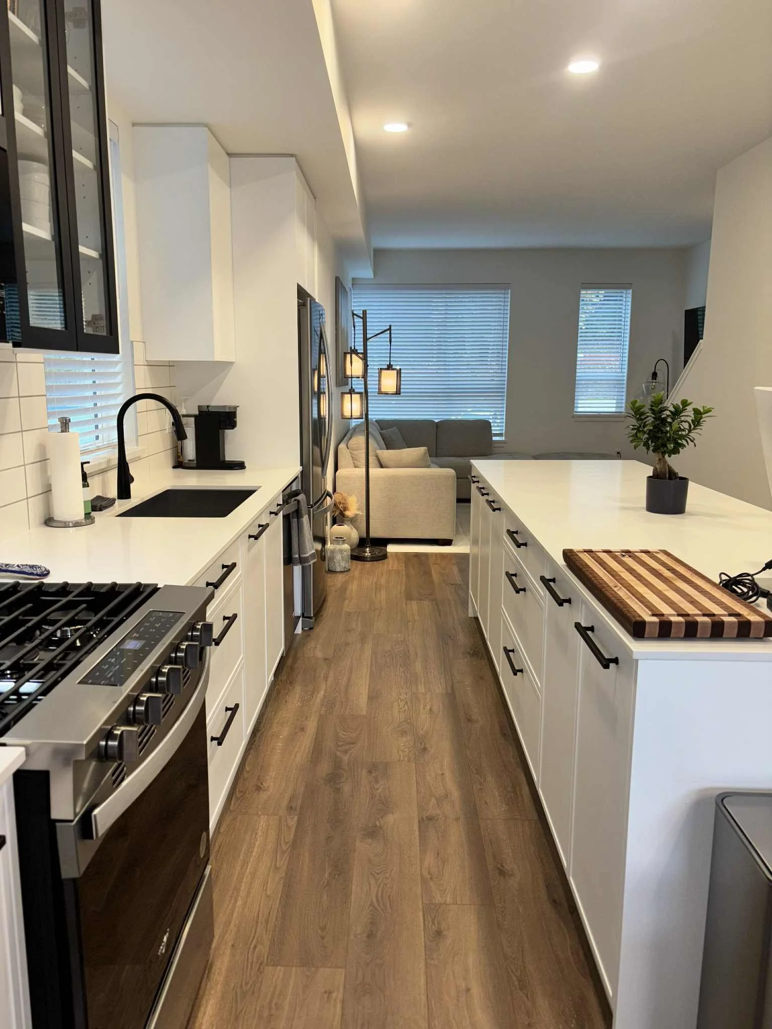 A modern kitchen with white cabinets, black handles, a black stove, and a white countertop. The kitchen opens into a living room with a beige sofa, large windows with blinds, and a standing lamp. There is a cutting board on the countertop, and a small potted plant.