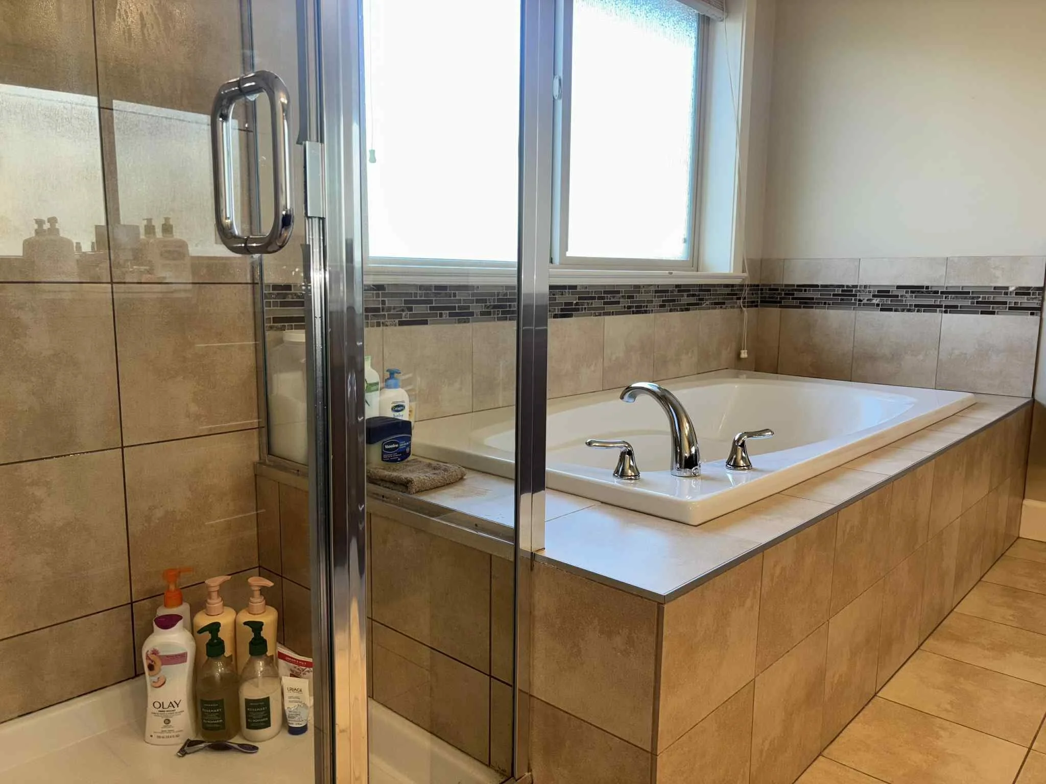 Bathroom with tiled walls and floor, a bathtub with chrome faucet and handles, a large window above the bathtub, and a separate shower stall with bottles of lotion and soap on the floor.