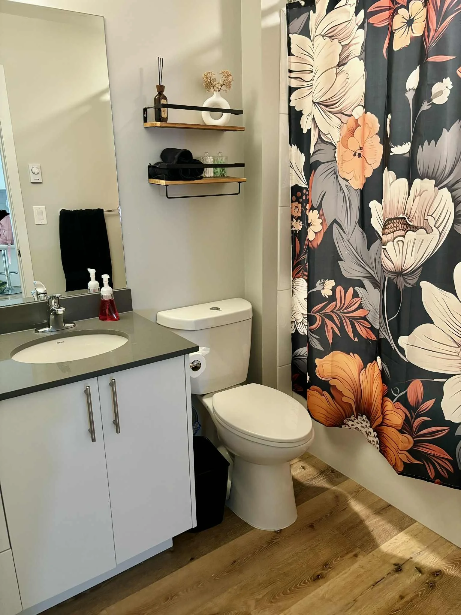 Bathroom with white vanity, gray countertop, toilet, floral shower curtain, wall shelves with decorative items, black towel, and wooden floor.