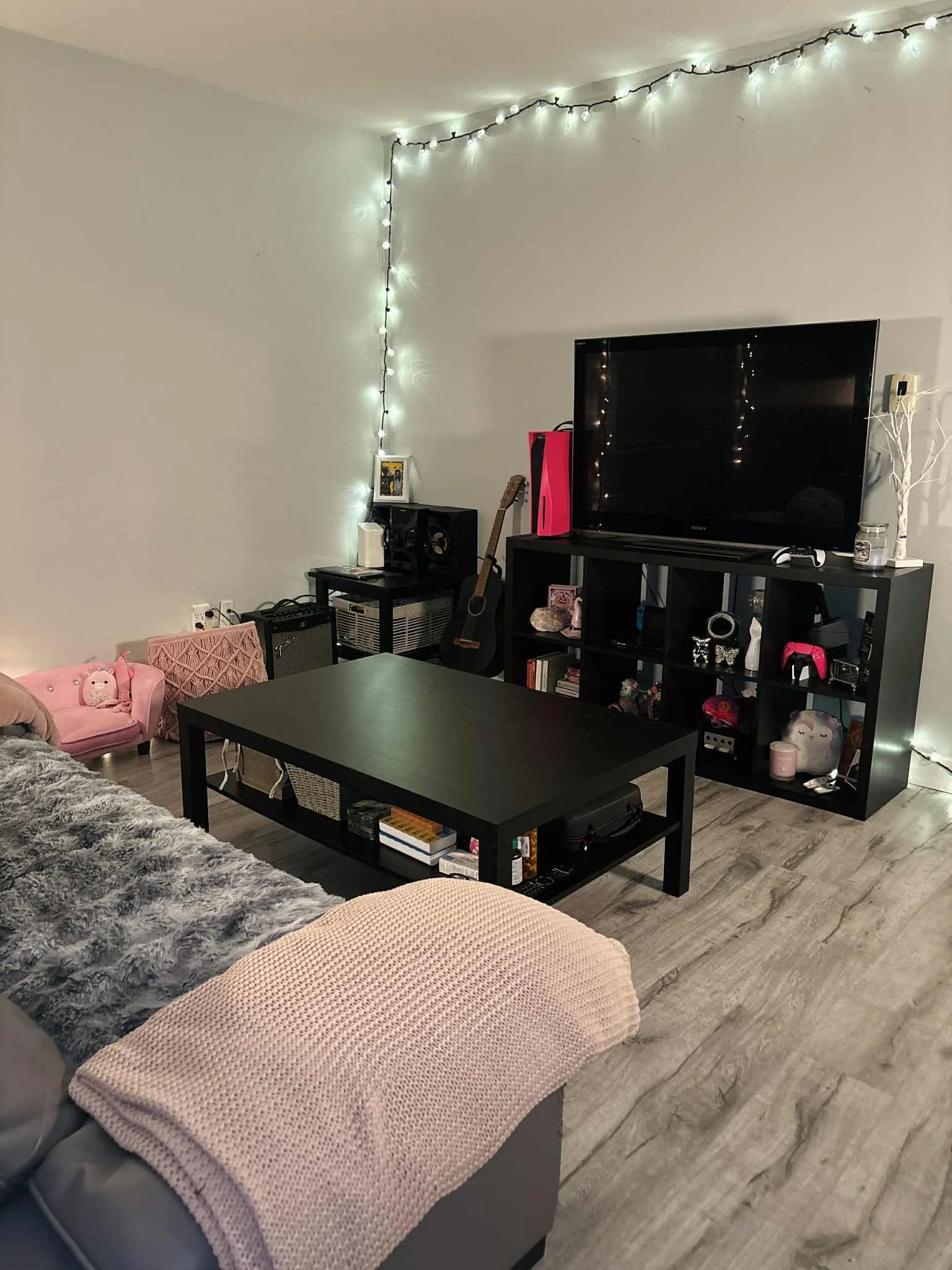 Living room with string lights on the ceiling, a black TV stand with various items including a guitar, pink gaming console, and decorations. A pink child's chair, a faux fur blanket on a gray couch, a black coffee table with books, and a bookshelf wi