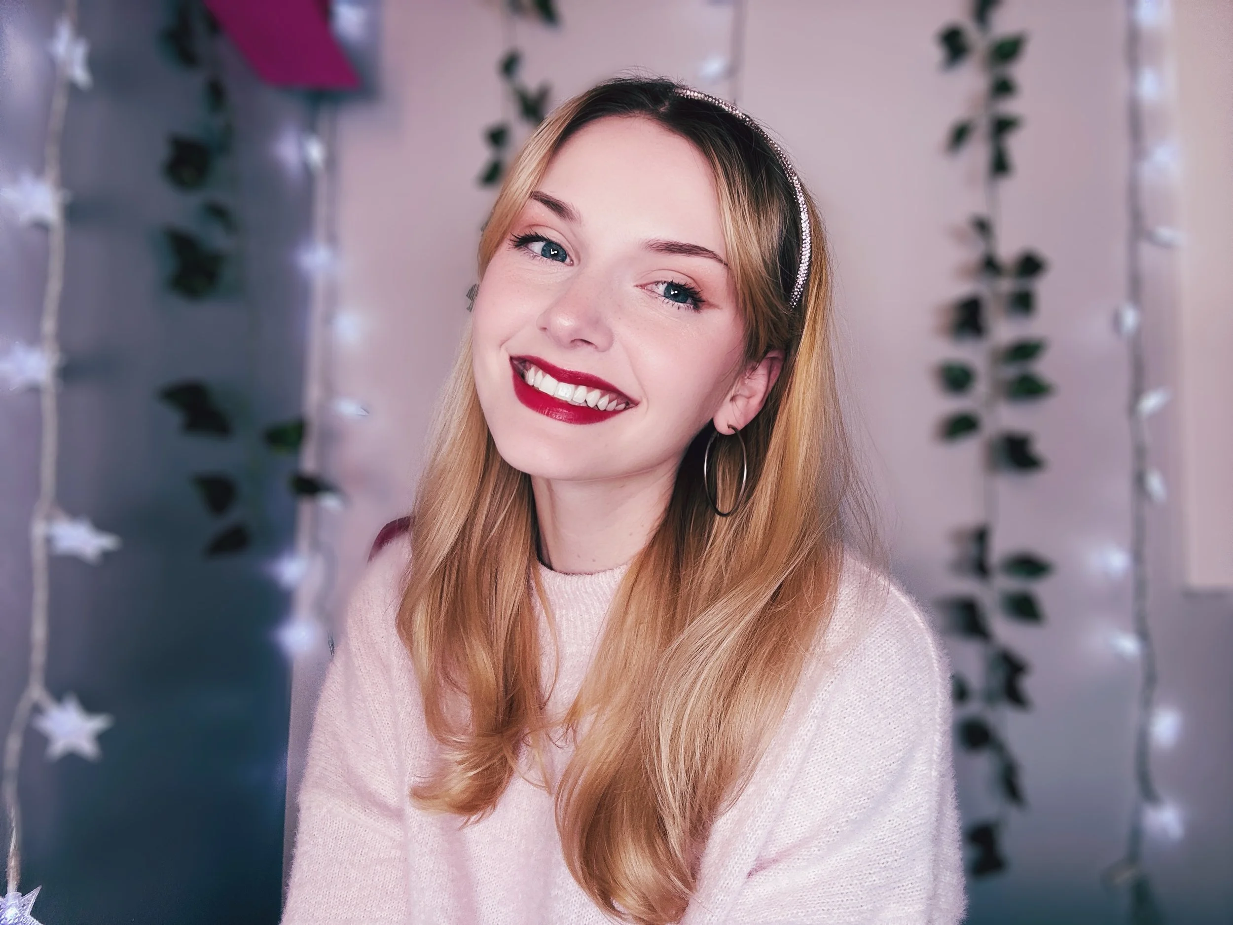 A smiling woman with blonde hair, red lipstick, and hoop earrings posing in front of a background with hanging star-shaped ornaments and lights.