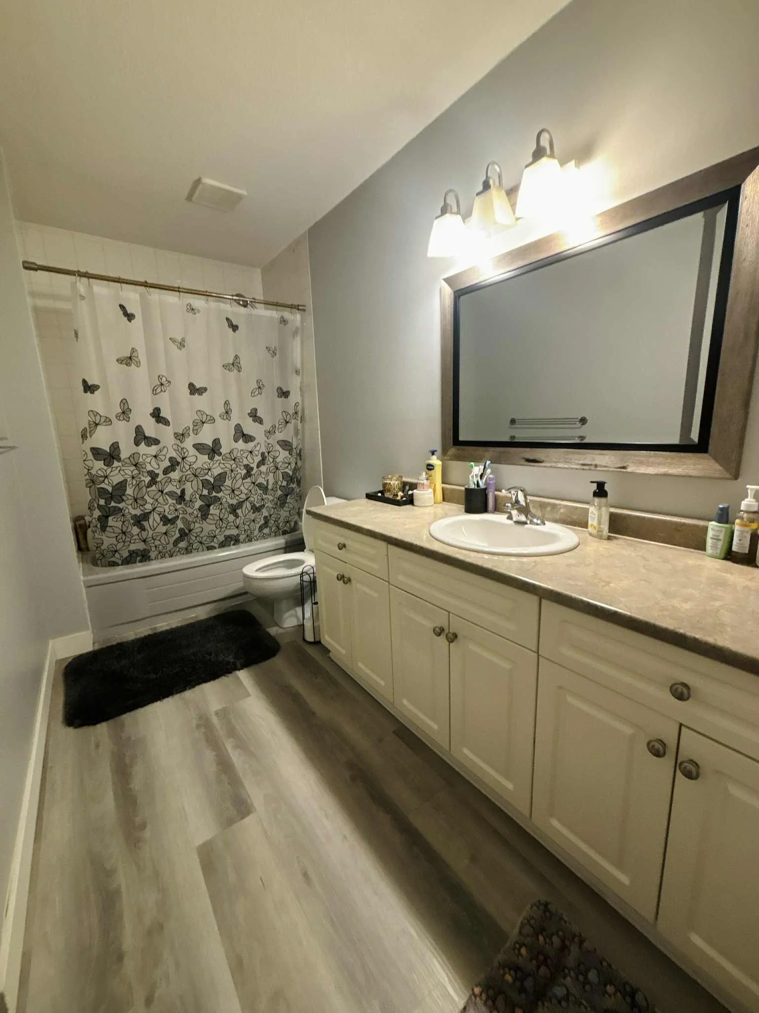 Bathroom with a beige vanity and large mirror, shower curtain with butterfly pattern, and toilet. Floor is wood-style laminate, black bath mat near the shower, and toiletries on the vanity.