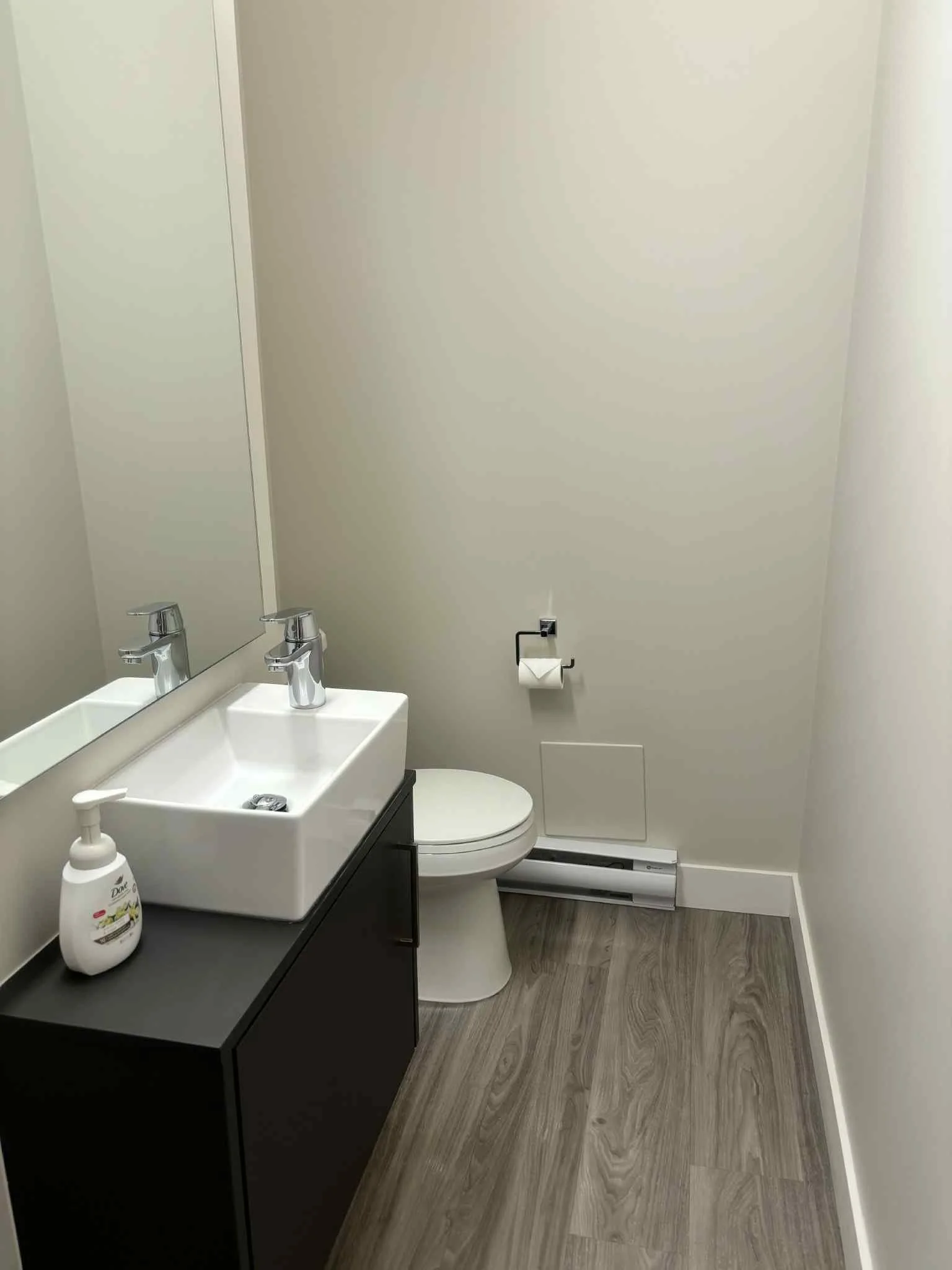 Small bathroom with a white sink on a black cabinet, a toilet, a mirror, and a dispenser of hand soap, with a wood-patterned floor and beige walls.