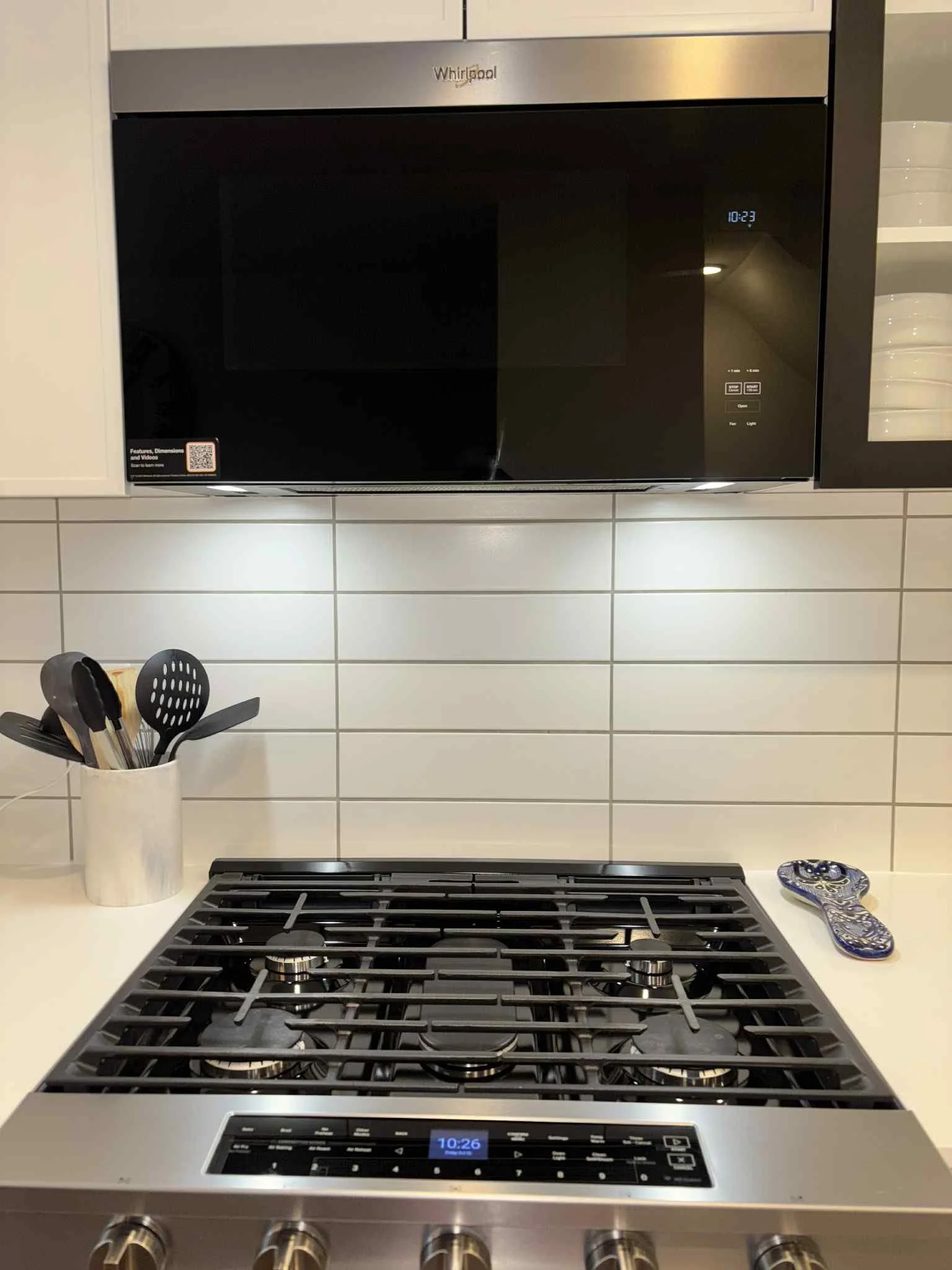 A modern kitchen with a black microwave over a gas stove, a utensil holder with black utensils, and a patterned pot holder on the counter.