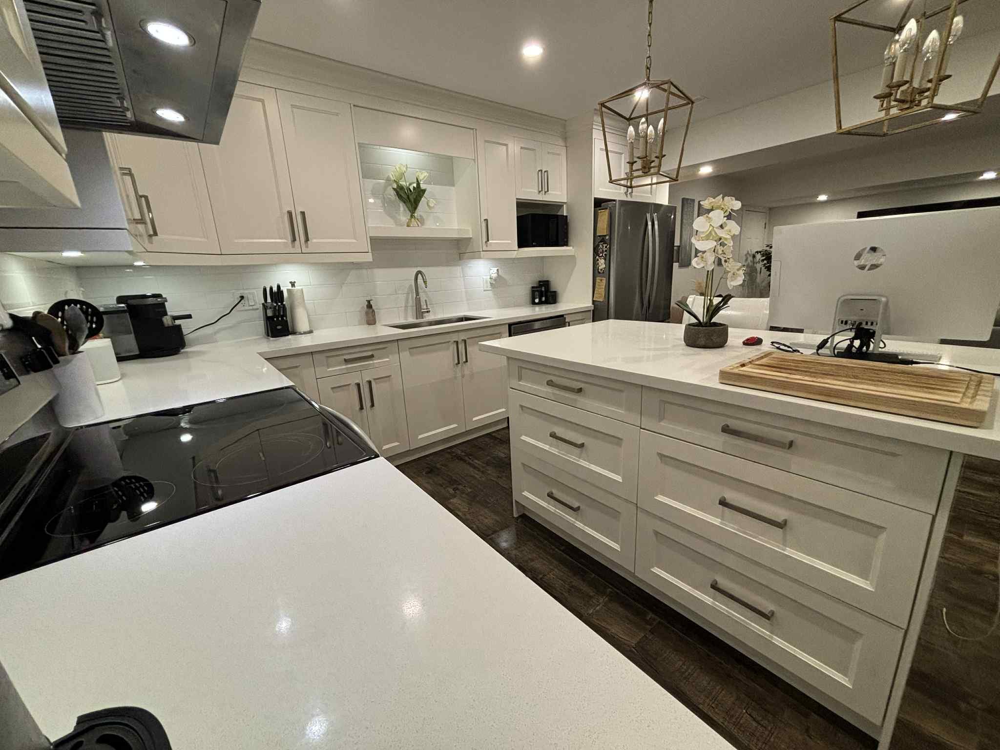 Modern kitchen with white cabinets, a white countertop, stainless steel appliances, a stove, a sink, a refrigerator, and decorative plants. There are pendant lights hanging from the ceiling and various kitchen tools on the counters.