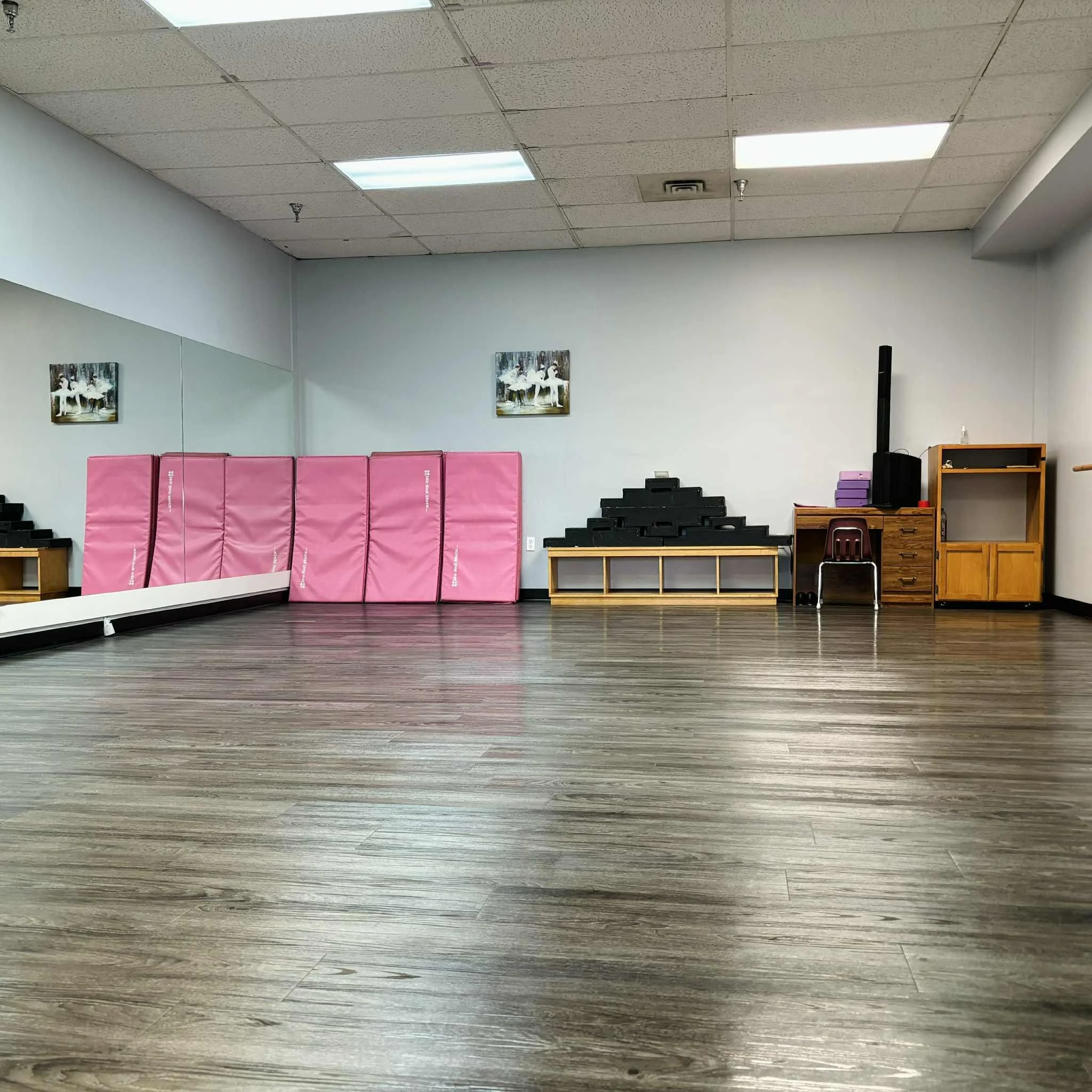 A spacious, empty dance or exercise studio with wood flooring, pink mats stacked against the wall, and shelves with black pyramid-shaped blocks, a small wooden desk with purple boxes, and a chair. There are two framed pictures of white swans on the w