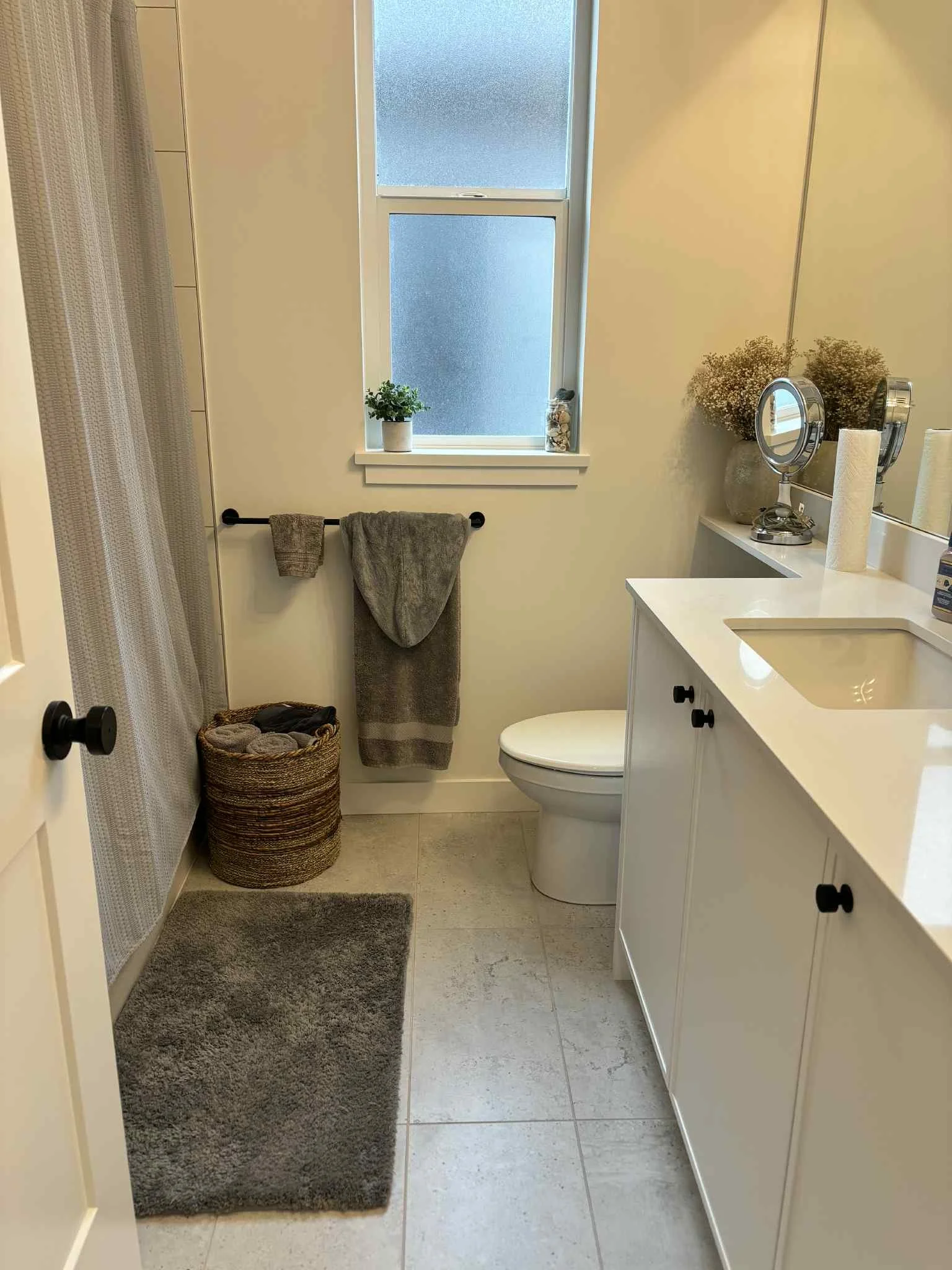 A small bathroom with a white vanity, a cream-colored countertop, and a sink. There is a mirror with two round magnifying mirrors attached. Two toilet paper rolls are on the counter, and a vase with dried flowers on the corner. A window with frosted glass lets in natural light, with a small potted plant and decorative shells on the windowsill. A towel rack with gray towels, a woven basket with rolled towels, a brown rug, and a shower curtain are also visible.