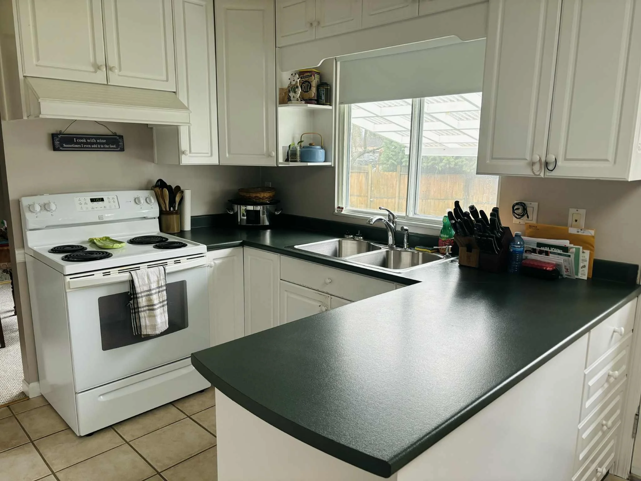 White kitchen with black countertops, stove, double sink, cabinetry, and window, with various kitchen items on the counters.