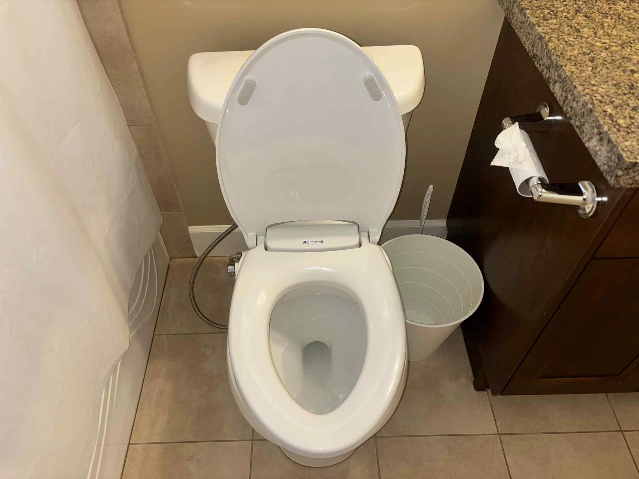 Toilet with the lid up next to a dark brown bathroom cabinet with a granite countertop, a white wastebasket, and a toilet paper roll.