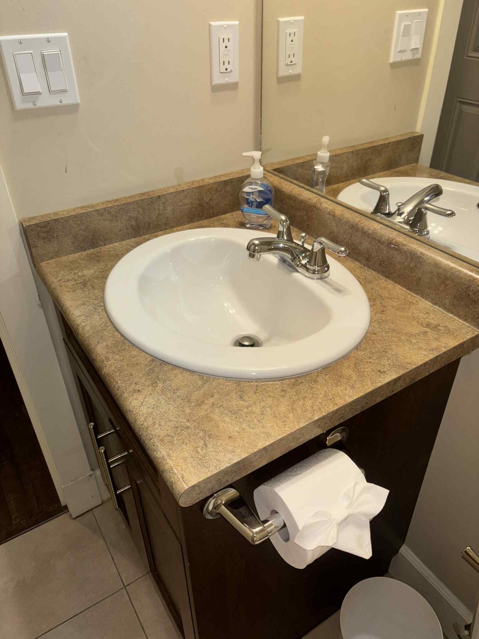 A bathroom vanity with a white sink, a chrome faucet, a mirror, and a soap dispenser. There are electrical outlets on the wall above the sink and a toilet paper roll mounted on the side of the cabinet with a tissue hanging from it.