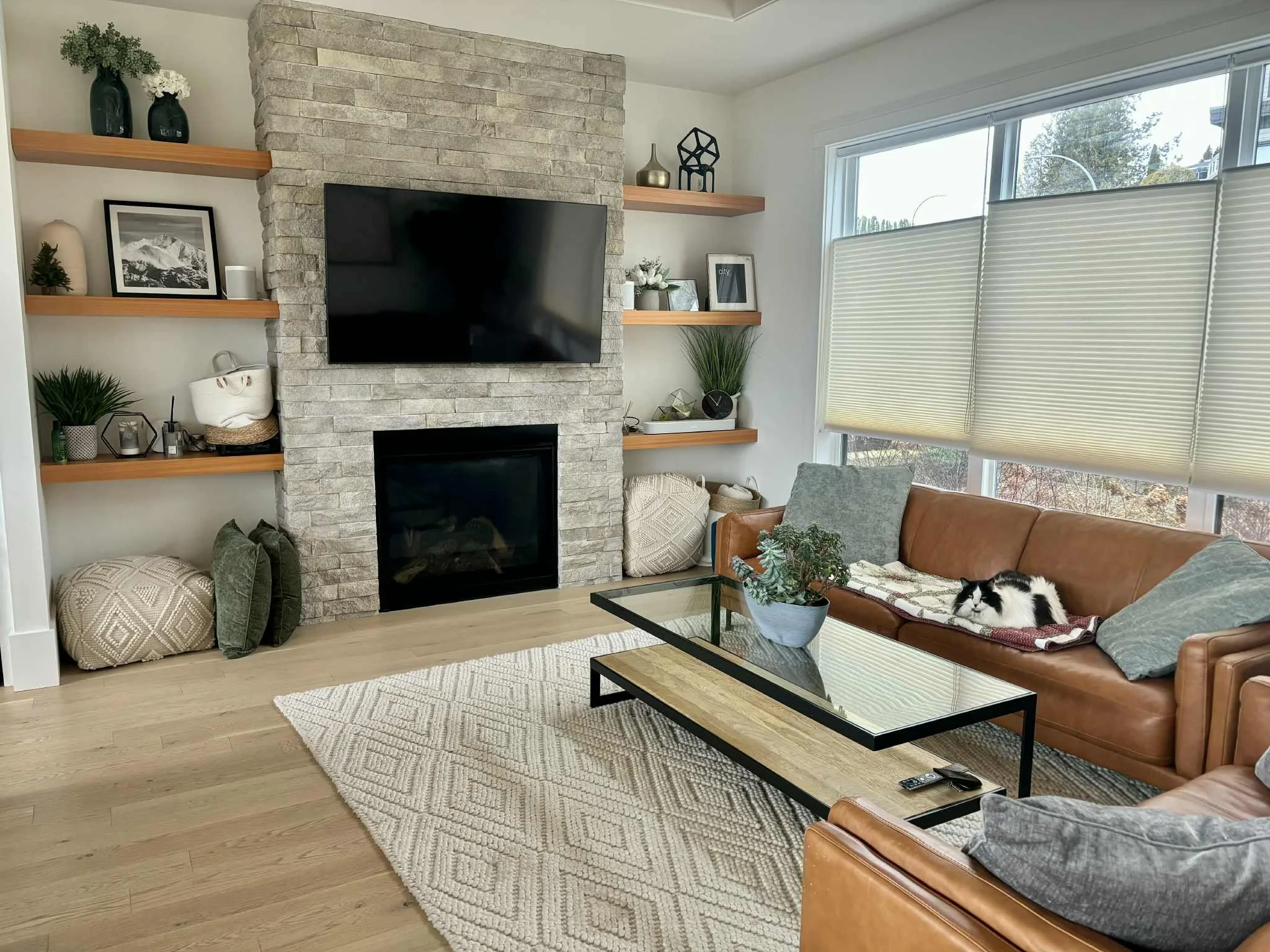 Cozy living room with a stone fireplace, wall-mounted TV, several open wooden shelves with decorative items, a tan leather sofa with pillows and a cat lying on it, a glass coffee table with a plant, and large front windows with blinds.
