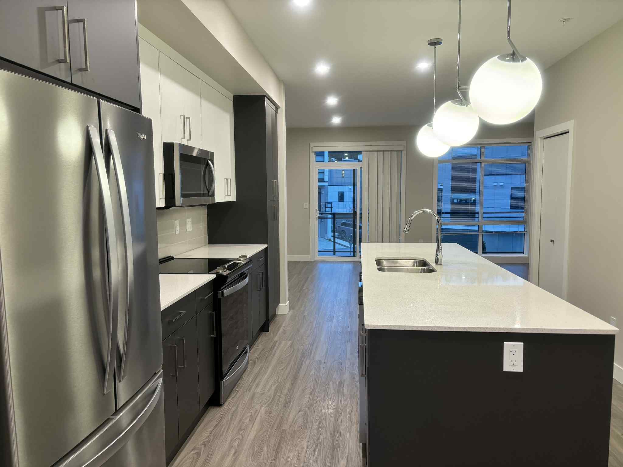 Modern kitchen with stainless steel appliances, white and dark cabinets, a large white island with a sink, pendant lights, and a view of an apartment balcony through sliding glass doors.