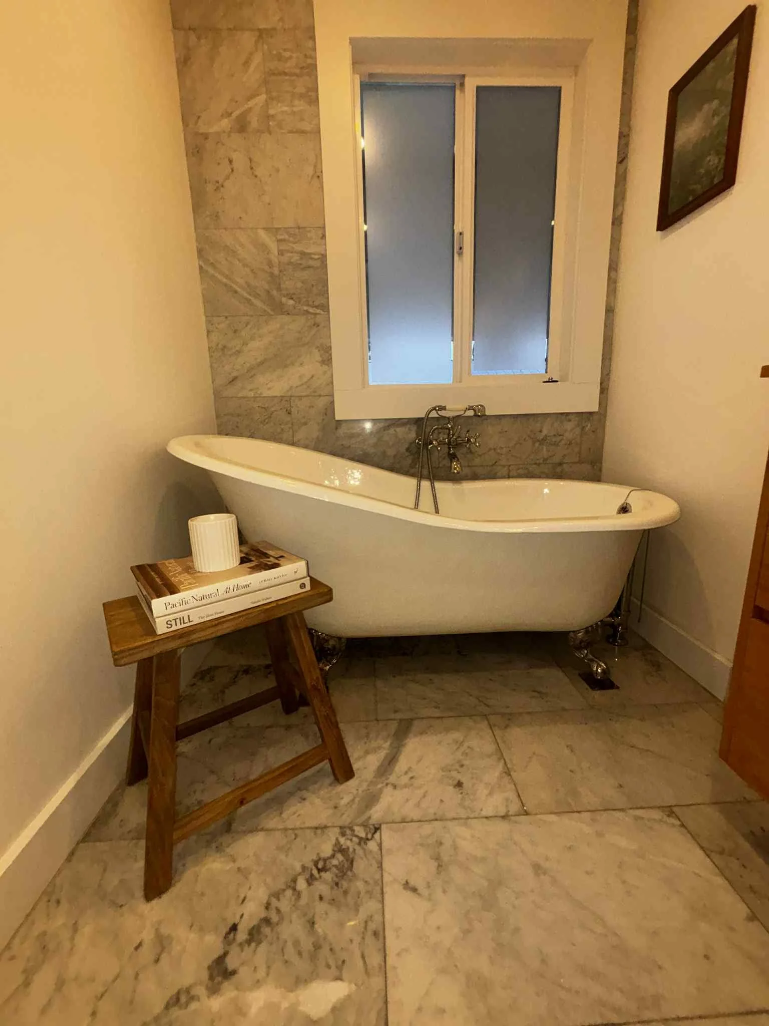 A bathroom with a vintage clawfoot bathtub positioned against a window and wall. There is a wooden bench with stacked books and a cup on it next to the tub. The floor and part of the wall are made of light-colored marble tiles, and a framed picture h