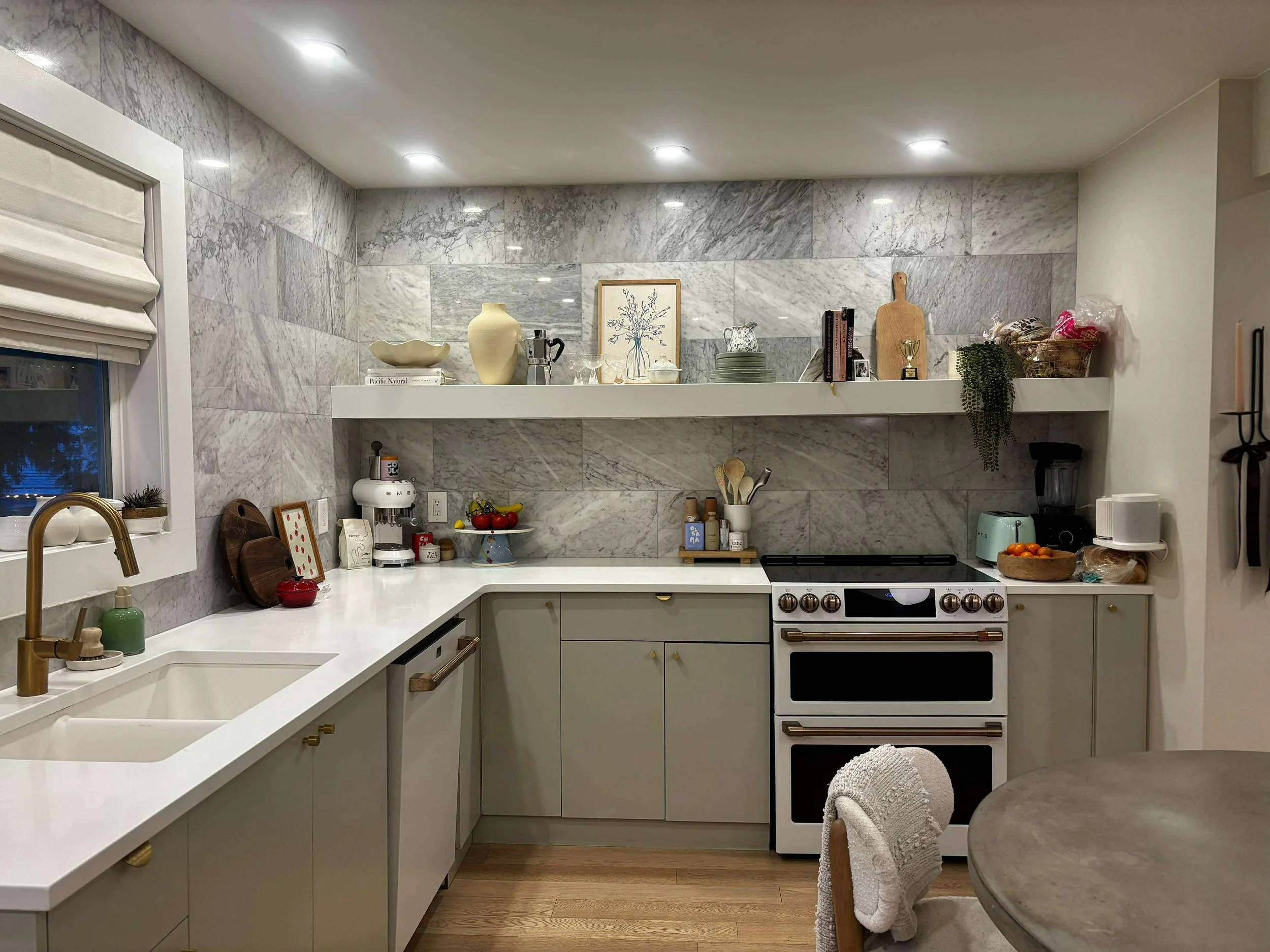 A modern kitchen with marble backsplash, white cabinets, a double sink with gold faucet, and various kitchen appliances and decor on the countertops and shelving.