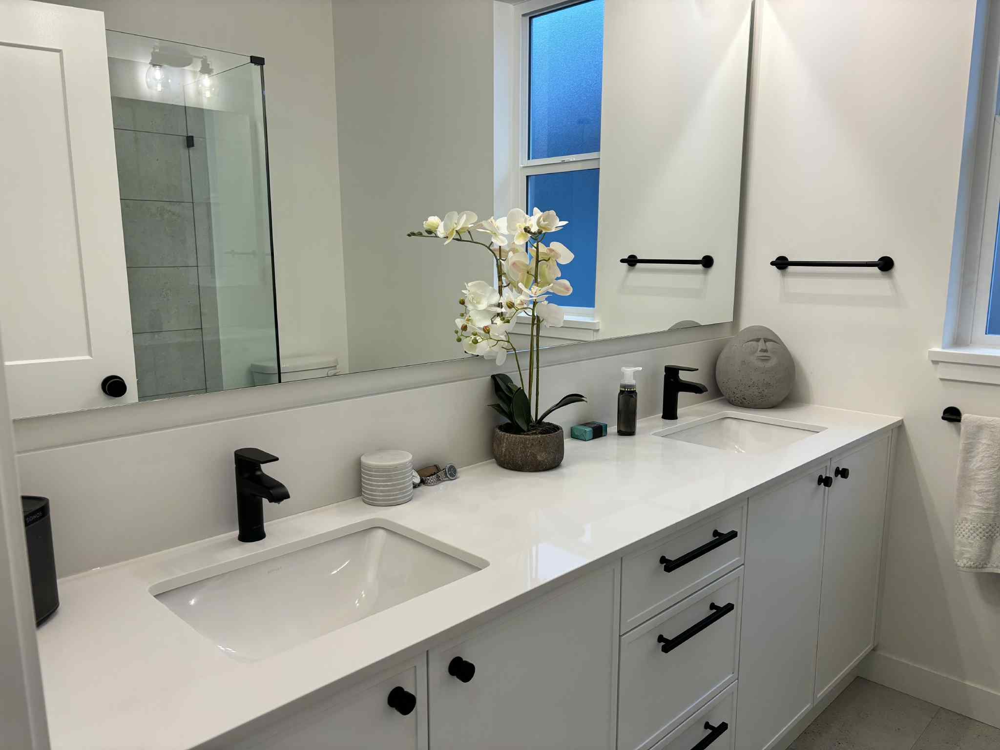 Modern bathroom with white vanity, black fixtures, large mirror, potted orchid, decorative stone sculpture, and a shower with glass door in background.
