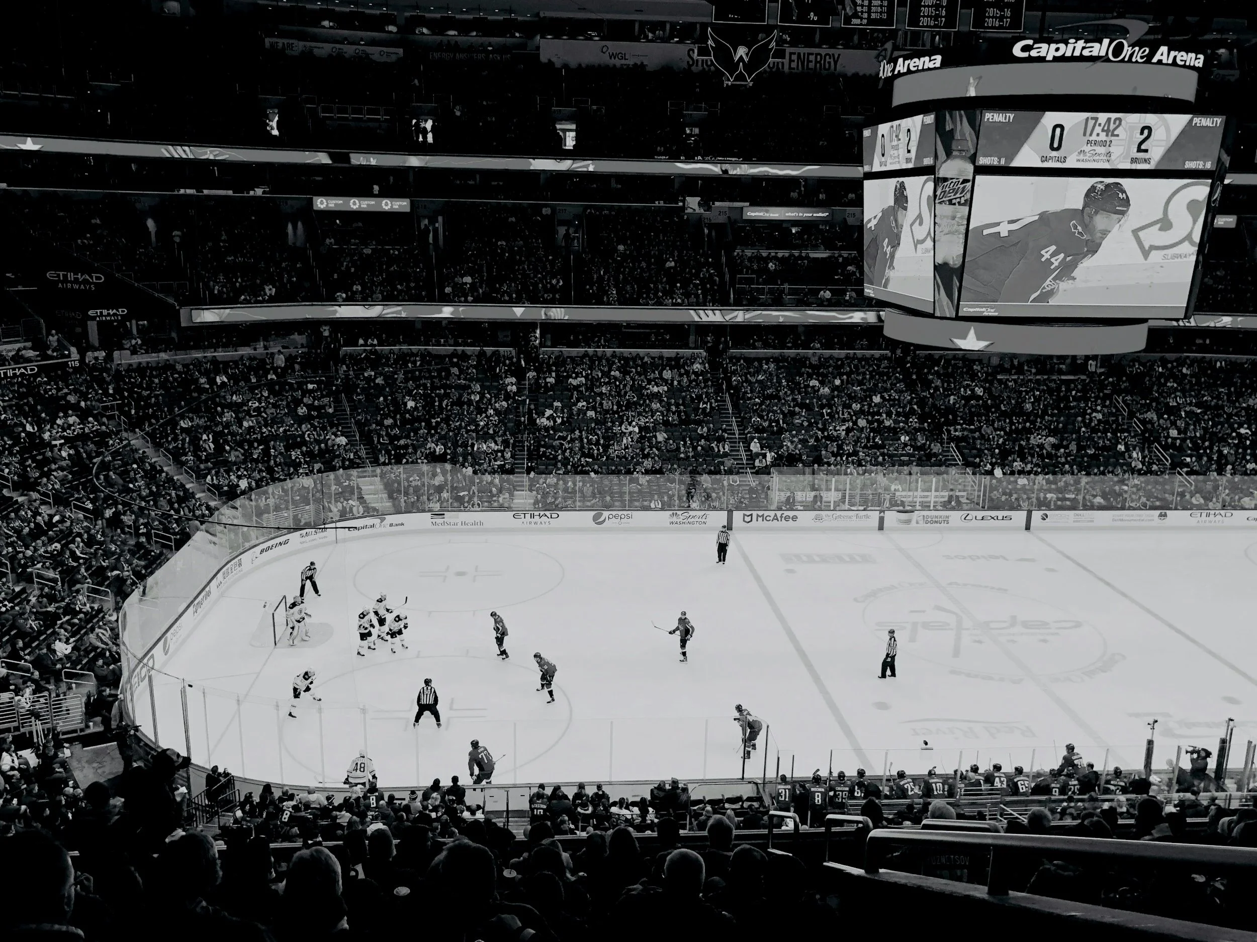Black and white photo of an ice hockey game in a large arena with fans in the seats. Players are on the ice, and officials are in striped shirts. A large digital scoreboard displays team scores, period, and game time.