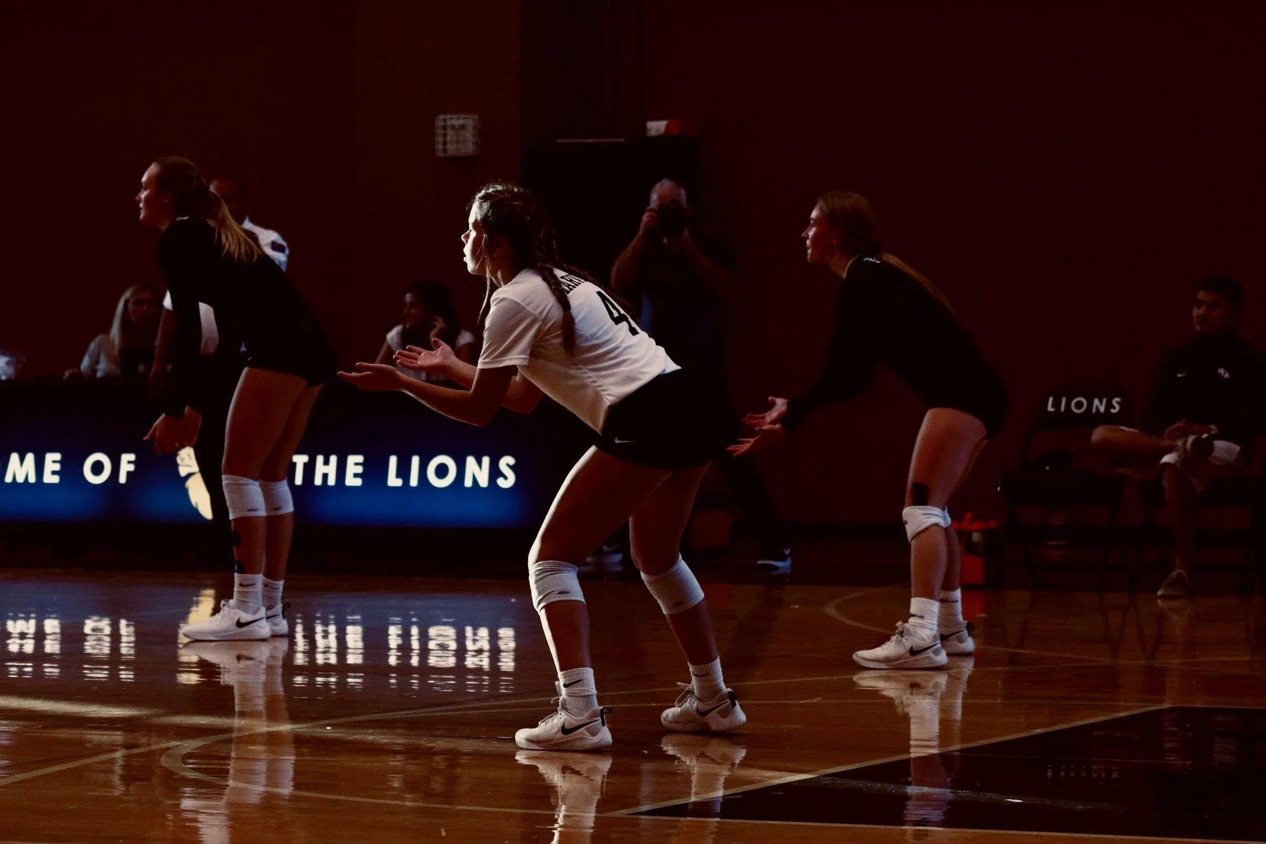 Volleyball players in black and white uniforms preparing to receive a serve on a court with a dark background and the text 'HOME OF THE LIONS' on an LED display.