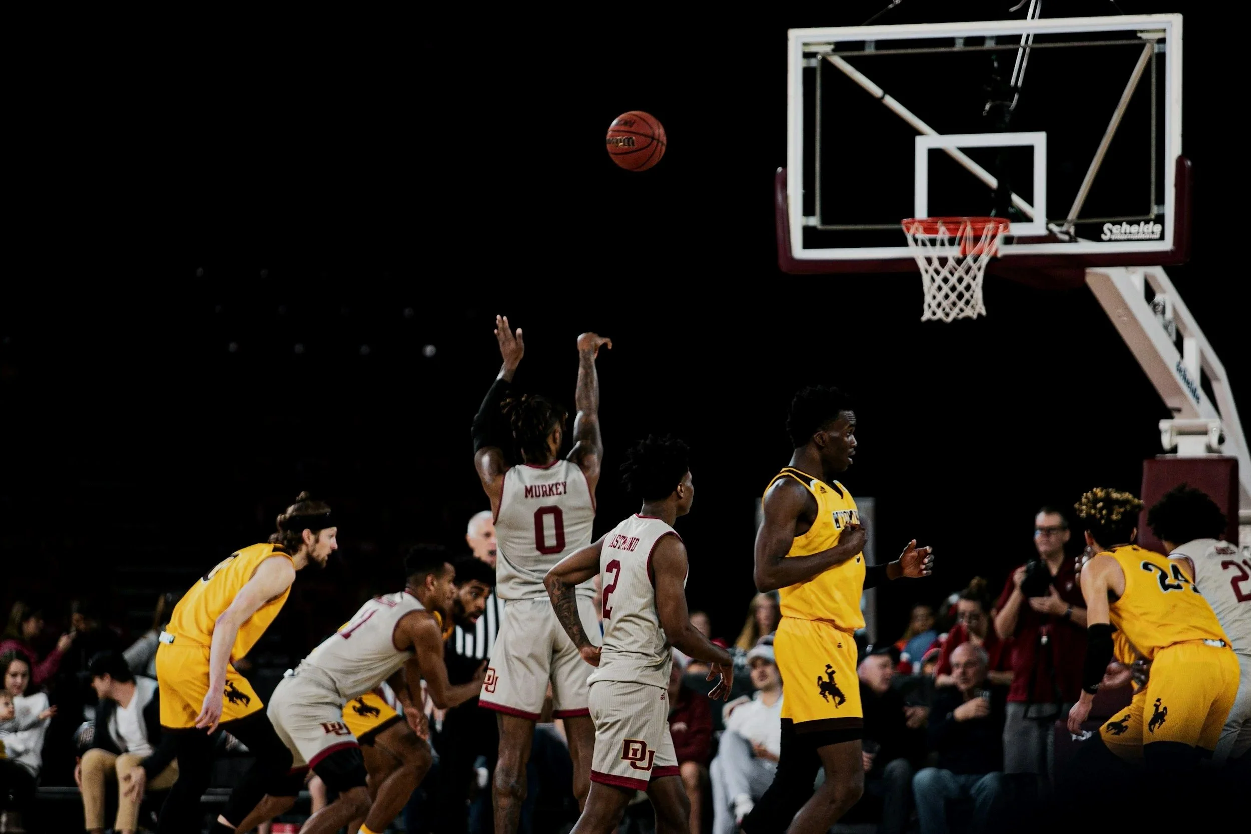 Basketball game with players from two teams, one in yellow uniforms and the other in white uniforms, on the court near the hoop, with spectators watching.