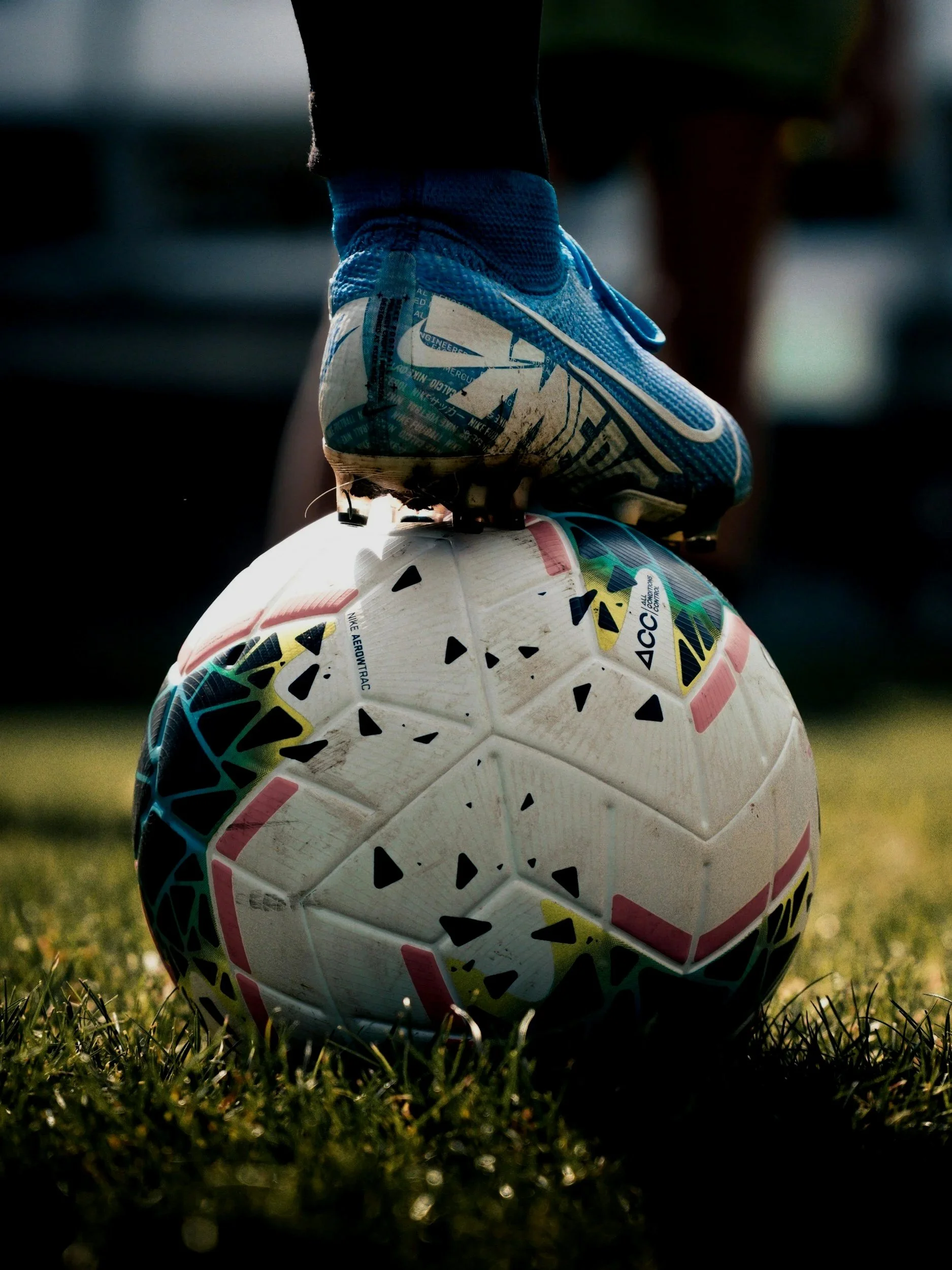 Close-up of a soccer ball on grass with a person wearing a blue athletic shoe and black socks standing on top of it.