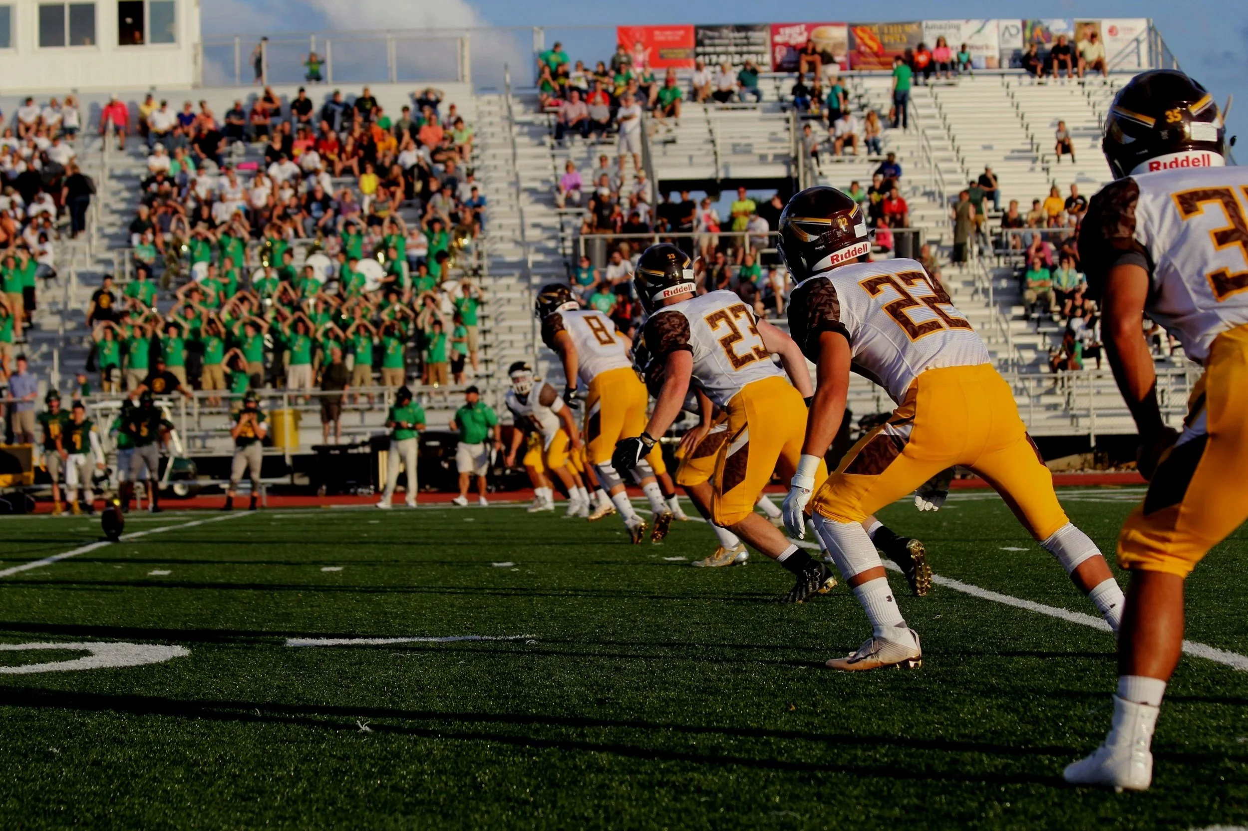 American football players in white and yellow uniforms lined up on the field, preparing for a play with spectators in the stands behind them.