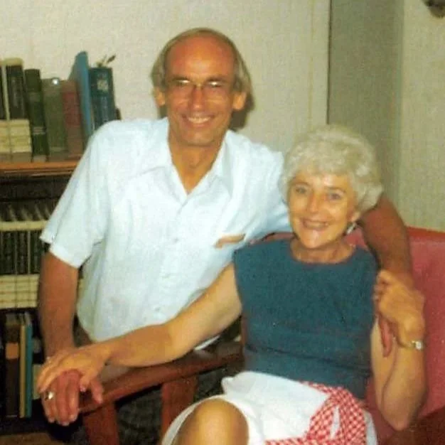 A smiling elderly woman sitting in a chair with a man standing beside her, holding her hand. The man is wearing glasses and a white shirt, and the woman has white hair and is dressed in a blue top with a white apron featuring a red checkered pocket.
