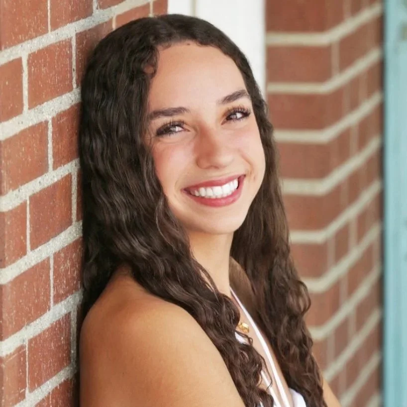 A young woman with long, curly brown hair and a bright smile, leaning against a brick wall outdoors.