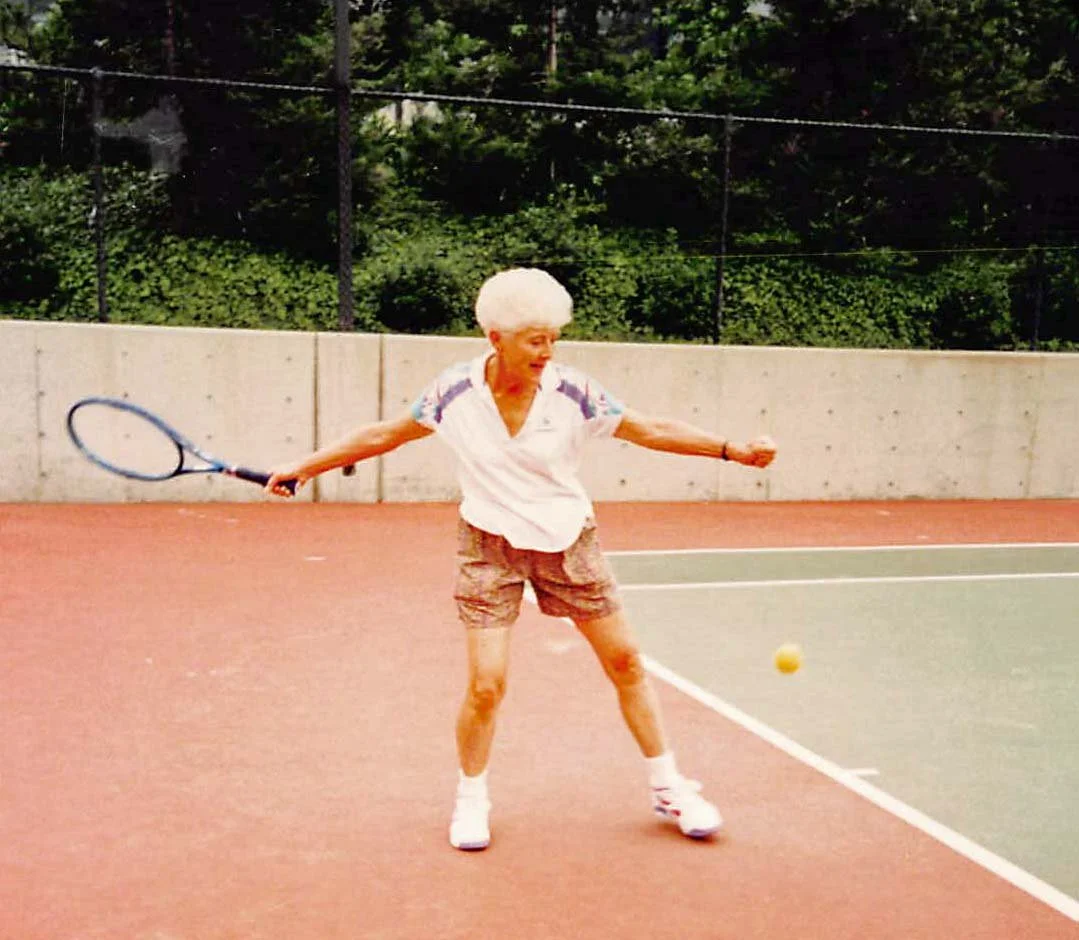 An elderly woman with white curly hair playing tennis on an outdoor court, swinging her racket toward a yellow tennis ball.