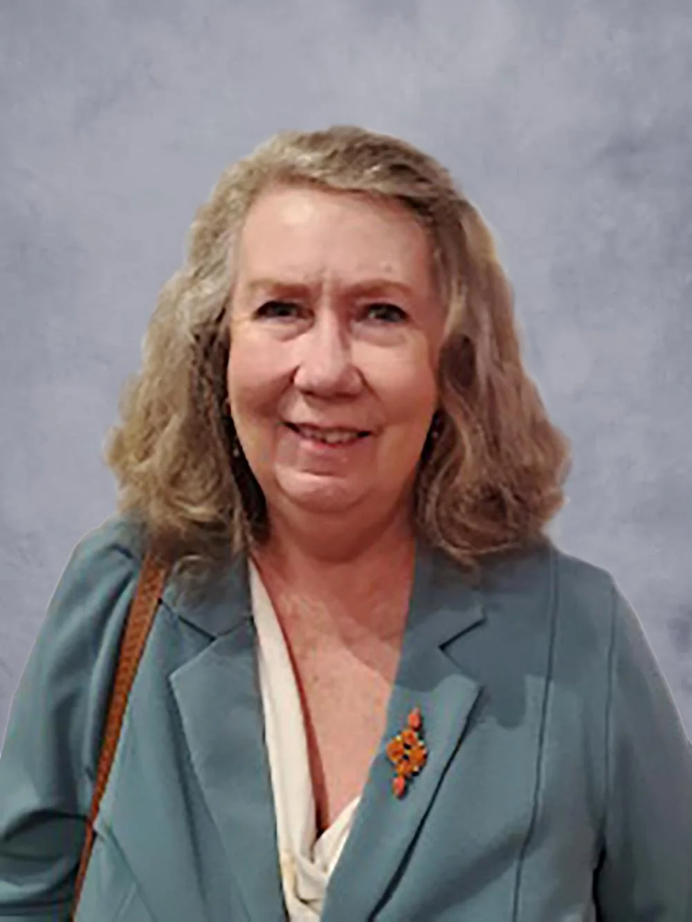 A smiling middle-aged woman with shoulder-length, wavy, light brown hair, wearing a teal blazer, a white top, and a brown strap from a bag or purse, standing indoors against a beige wall next to a wooden door or panel.