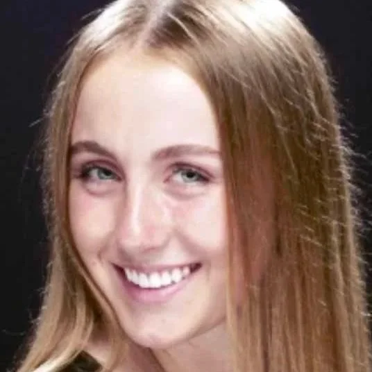 A young woman with long red hair smiling at the camera against a black background.