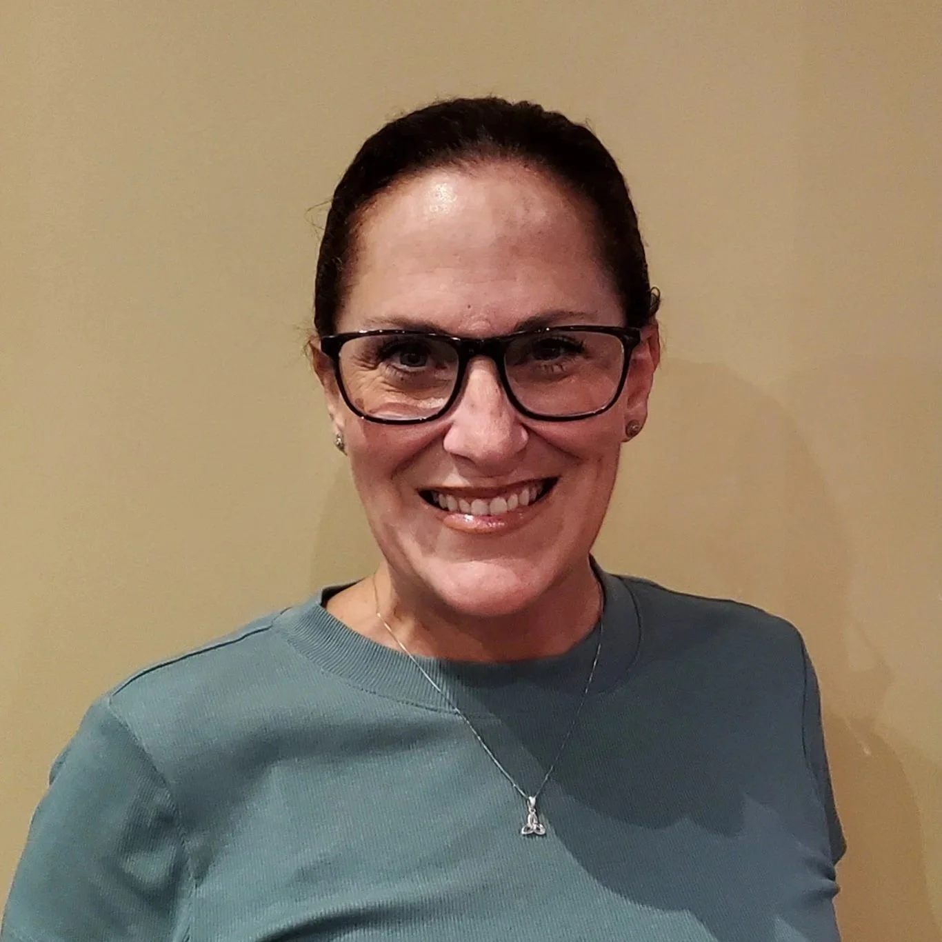 A woman with dark hair, black glasses, and a bright smile, wearing a gray shirt and a necklace, standing in front of a plain beige wall.