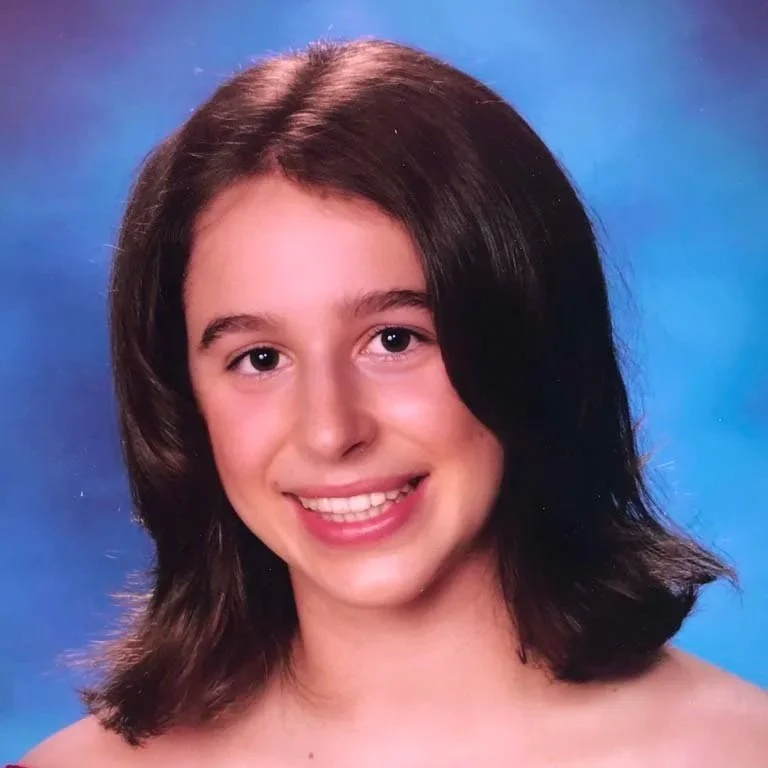 Smiling young girl with shoulder-length brown hair and light skin, posing against a blue background.