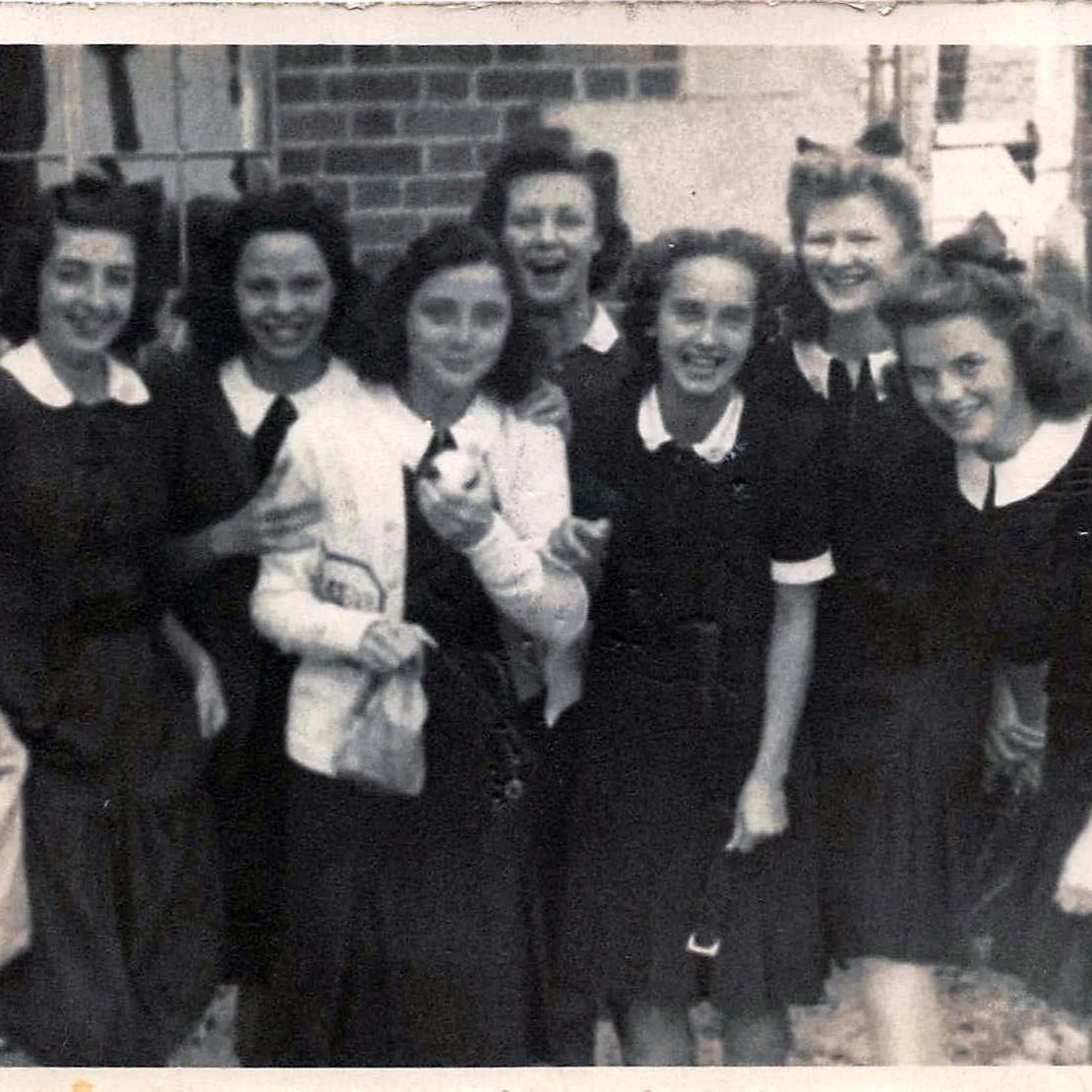 Black and white vintage photograph of seven women with curly hair, smiling and posing together outdoors in front of a brick wall.