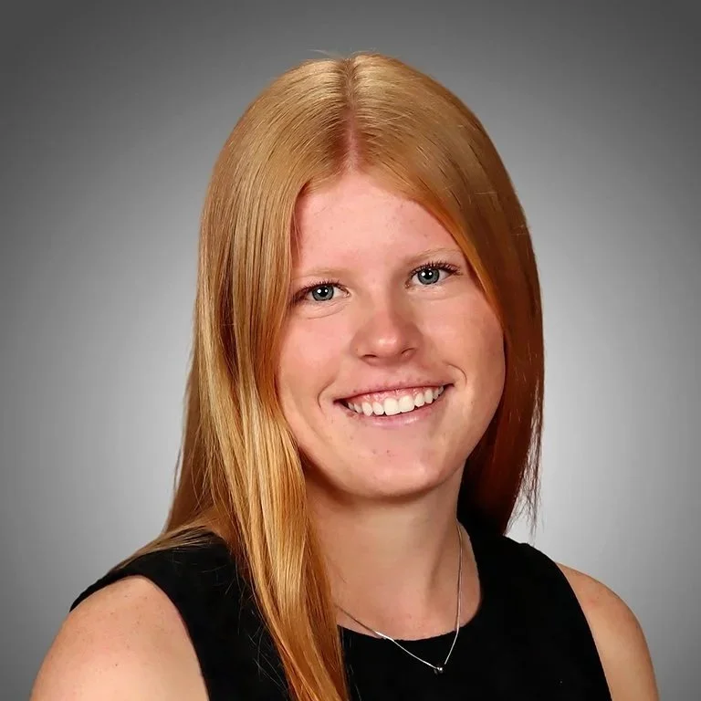 Young woman with long red hair, smiling, wearing a black sleeveless top, against a plain gray background.