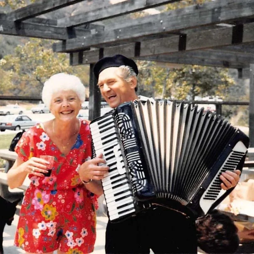 An elderly woman and a man playing an accordion outdoors, both smiling, with trees and parked cars in the background.