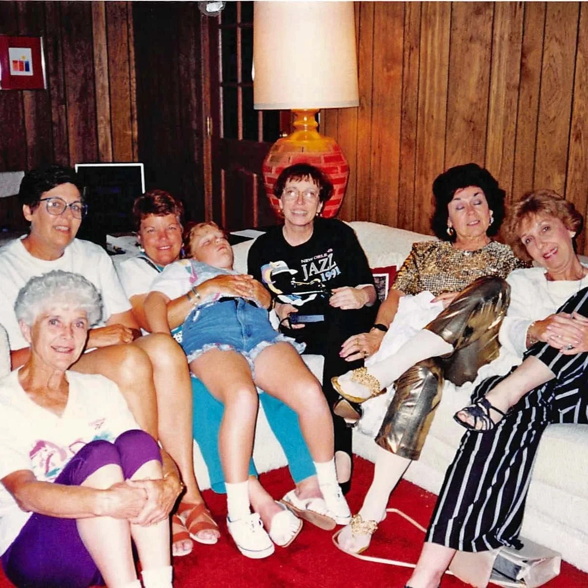 Seven women sitting and lying on sofas in a living room with wood-paneled walls, smiling and relaxing.