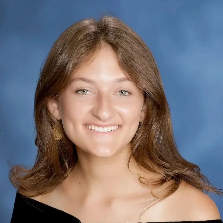 A smiling woman with long, wavy brown hair and light-colored eyes, wearing a black off-shoulder top and gold earrings, posed against a blue background.
