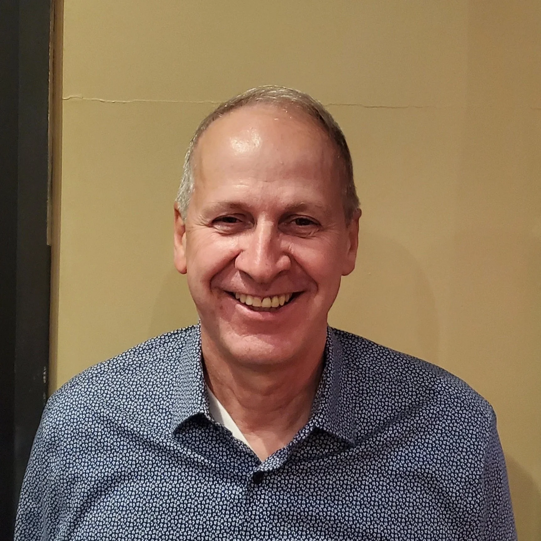 Smiling middle-aged man with short light-colored hair, wearing a dark patterned shirt, in front of a beige wall.