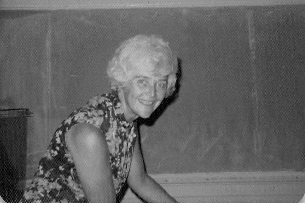 Black and white photo of an elderly woman with short, curly hair, smiling, wearing a sleeveless patterned dress, leaning forward.