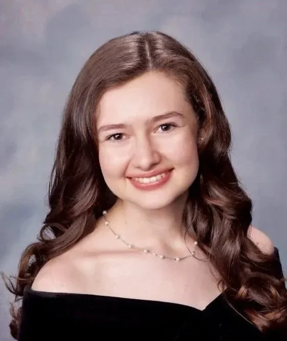 A young woman with long, wavy brown hair and light skin, smiling and wearing a black off-the-shoulder top with a delicate necklace, posing against a neutral background.