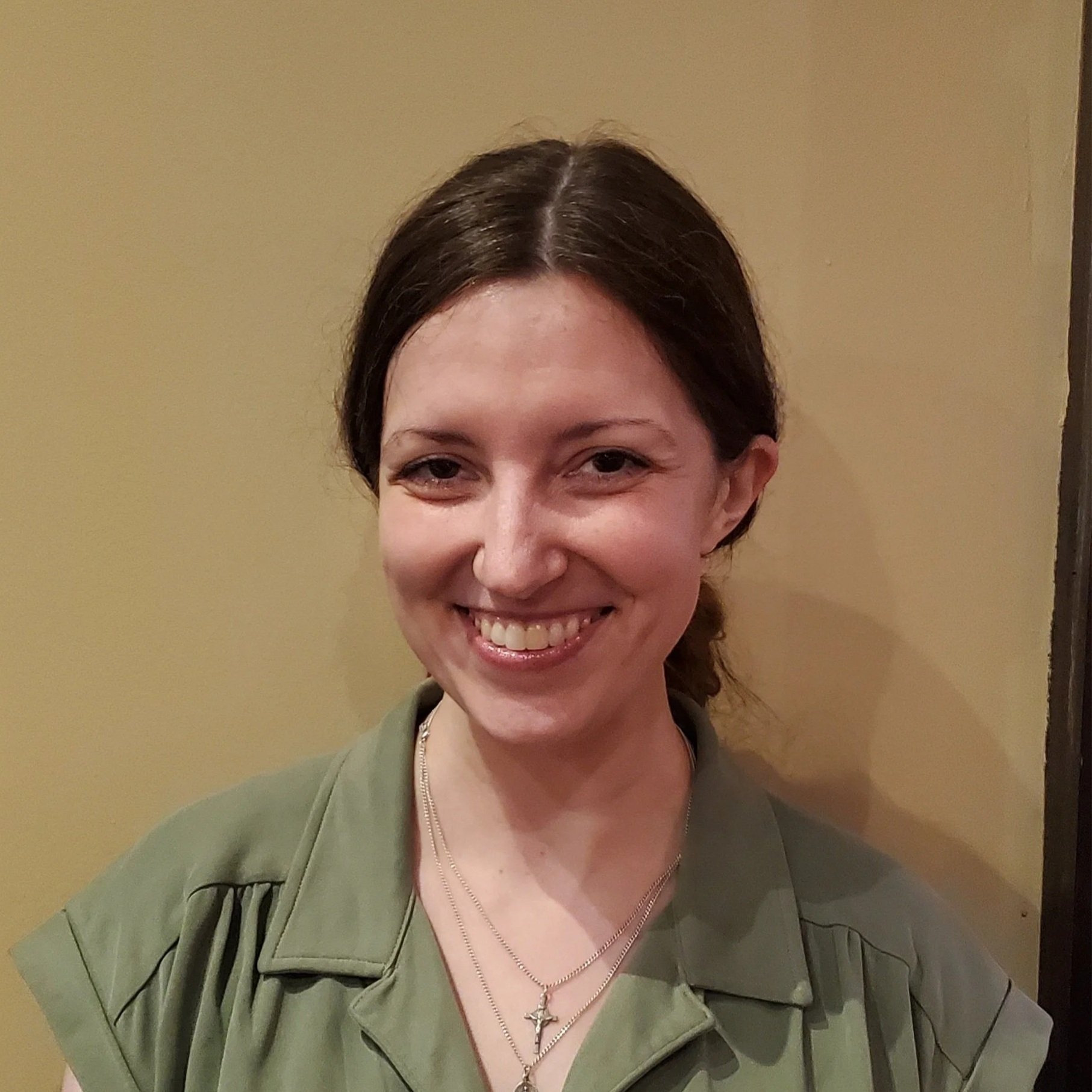A smiling woman with dark brown hair pulled back, wearing an olive-green shirt and layered necklaces with cross pendants, standing against a beige-colored wall.