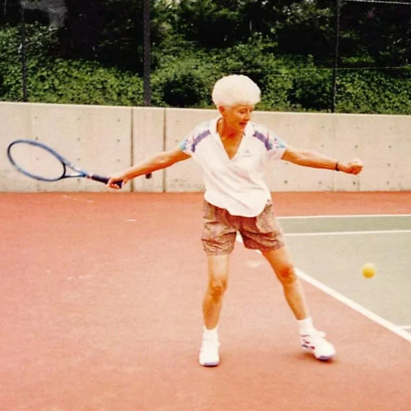 An elderly woman playing tennis on an outdoor court, preparing to hit a tennis ball with a racket.