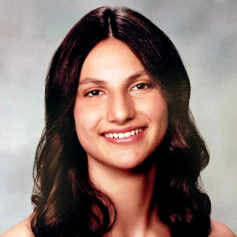 A young woman with long dark hair smiling at the camera against a plain light-colored background.