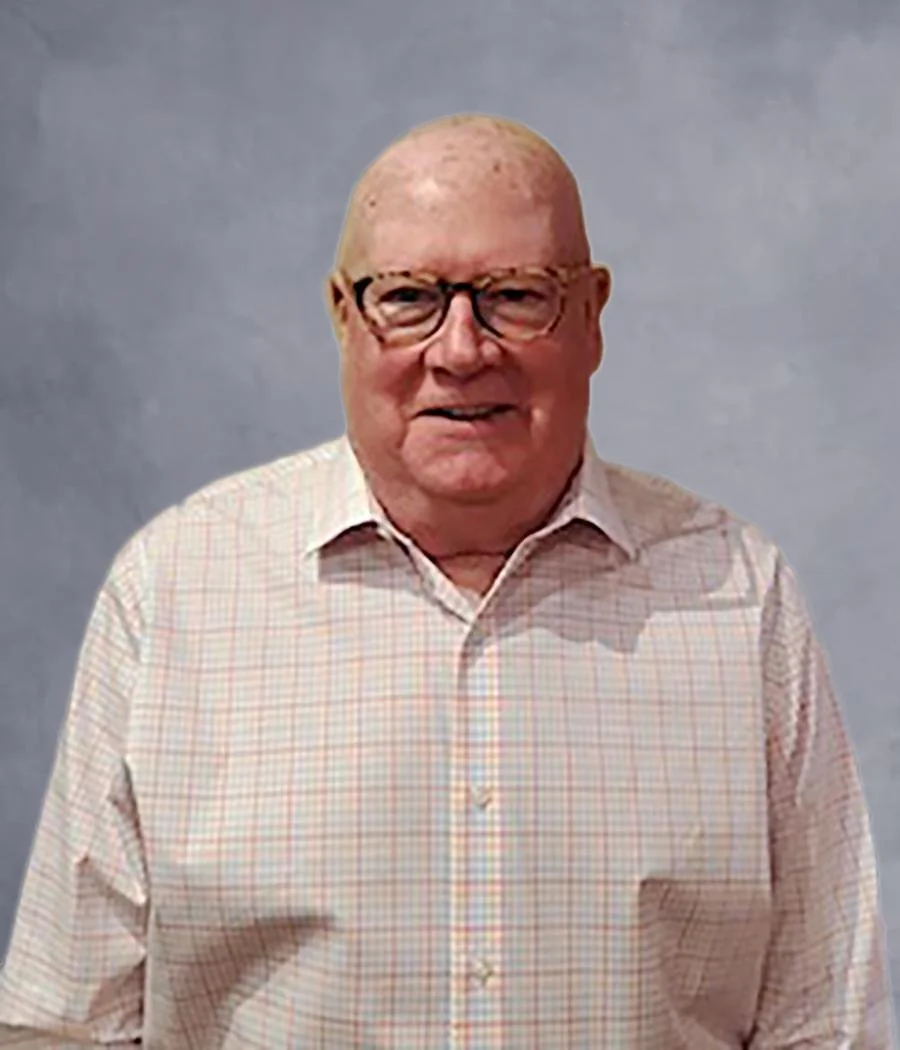 A man with glasses, short light hair, and a light checkered shirt standing against a beige wall.