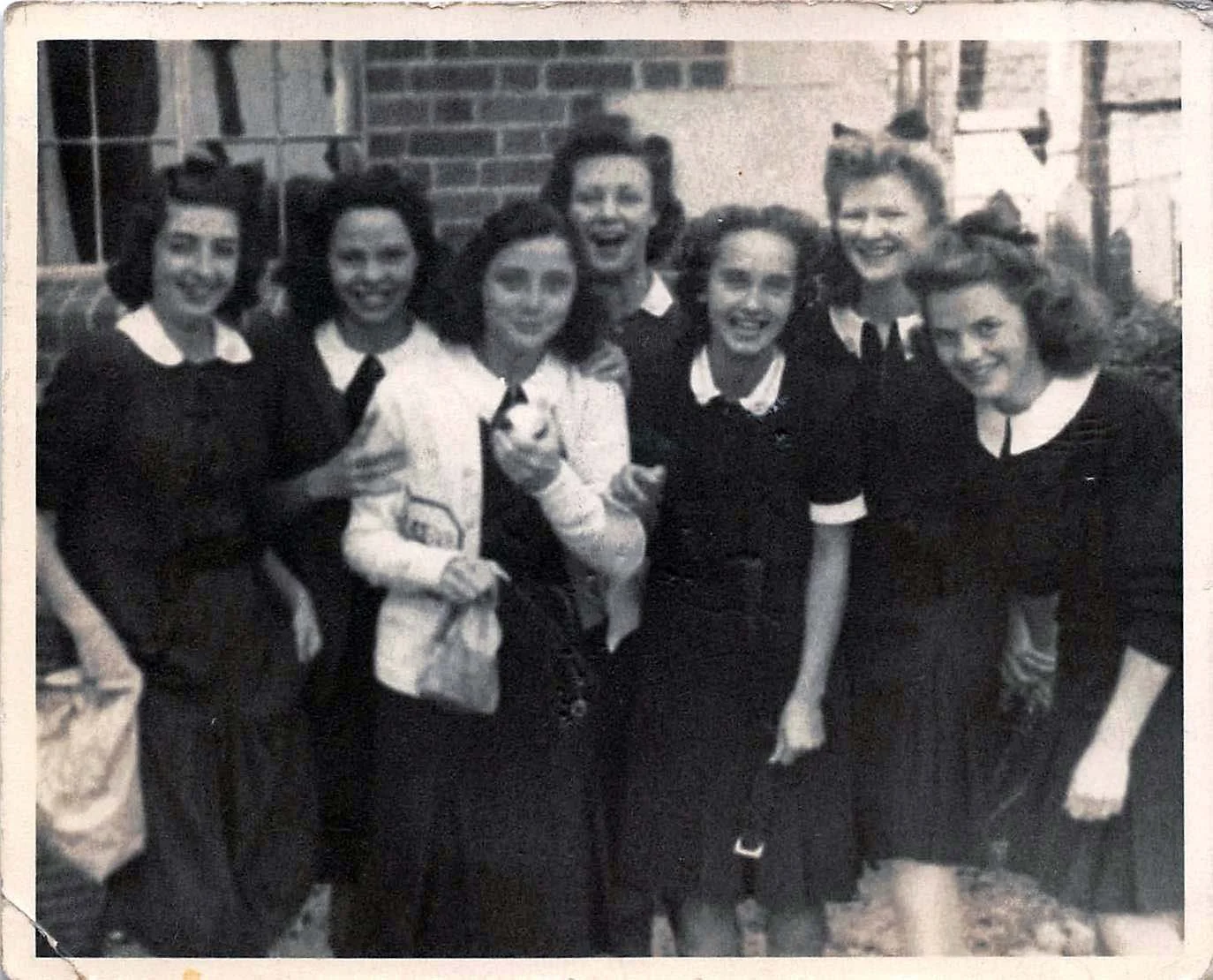 A black and white photo of seven women standing outdoors, dressed in similar dark tops with white collars, smiling and posing together.