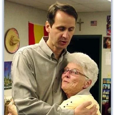A young man embraces an elderly woman in a room decorated with American flags and religious symbols.