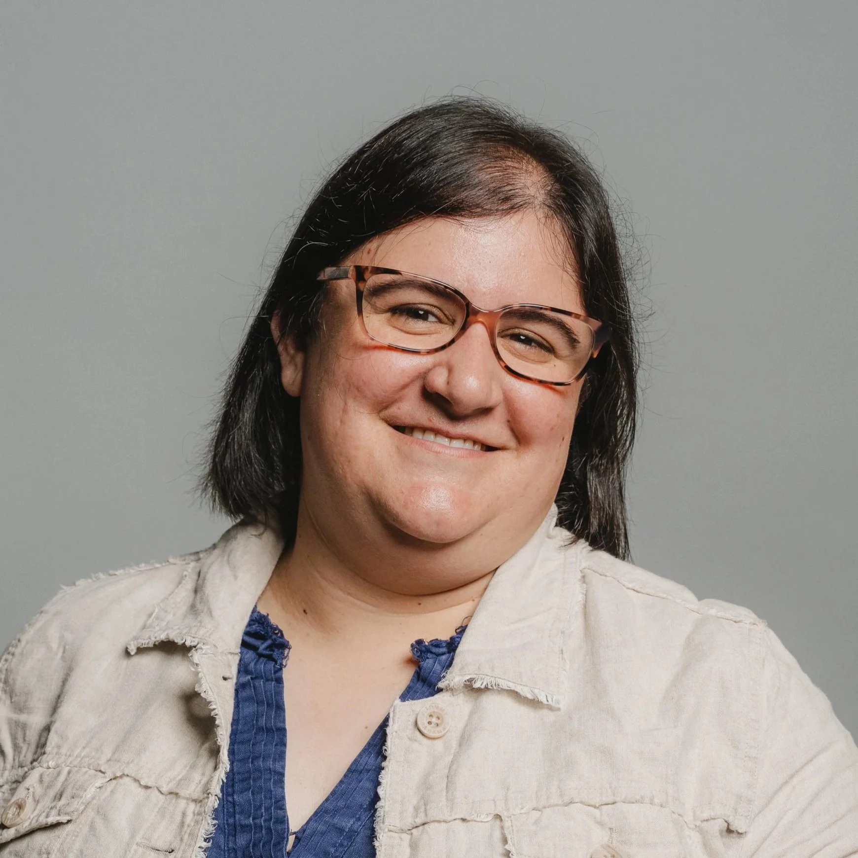A woman with shoulder-length dark hair, wearing glasses, smiling against a plain gray background.